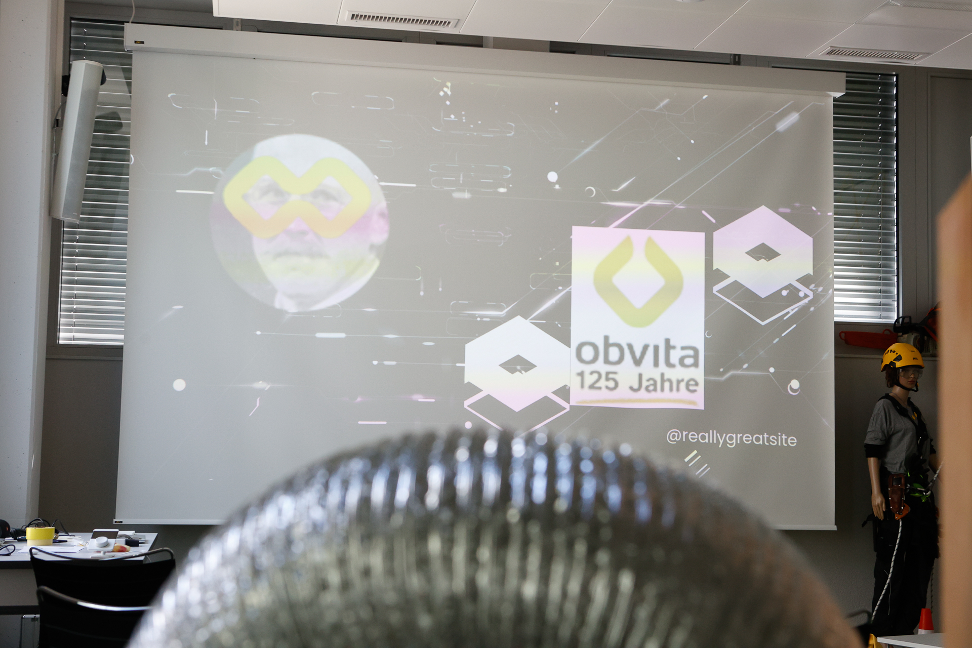 Auf einer Projektionsfläche sind die Worte „obvita 125 Jahre“ mit einem gelben Logo zu sehen. Rechts davon steht eine Person mit einem Helm. Im Vordergrund ist ein glänzendes, strukturiertes Objekt zu sehen, möglicherweise ein Lüftungskanal.