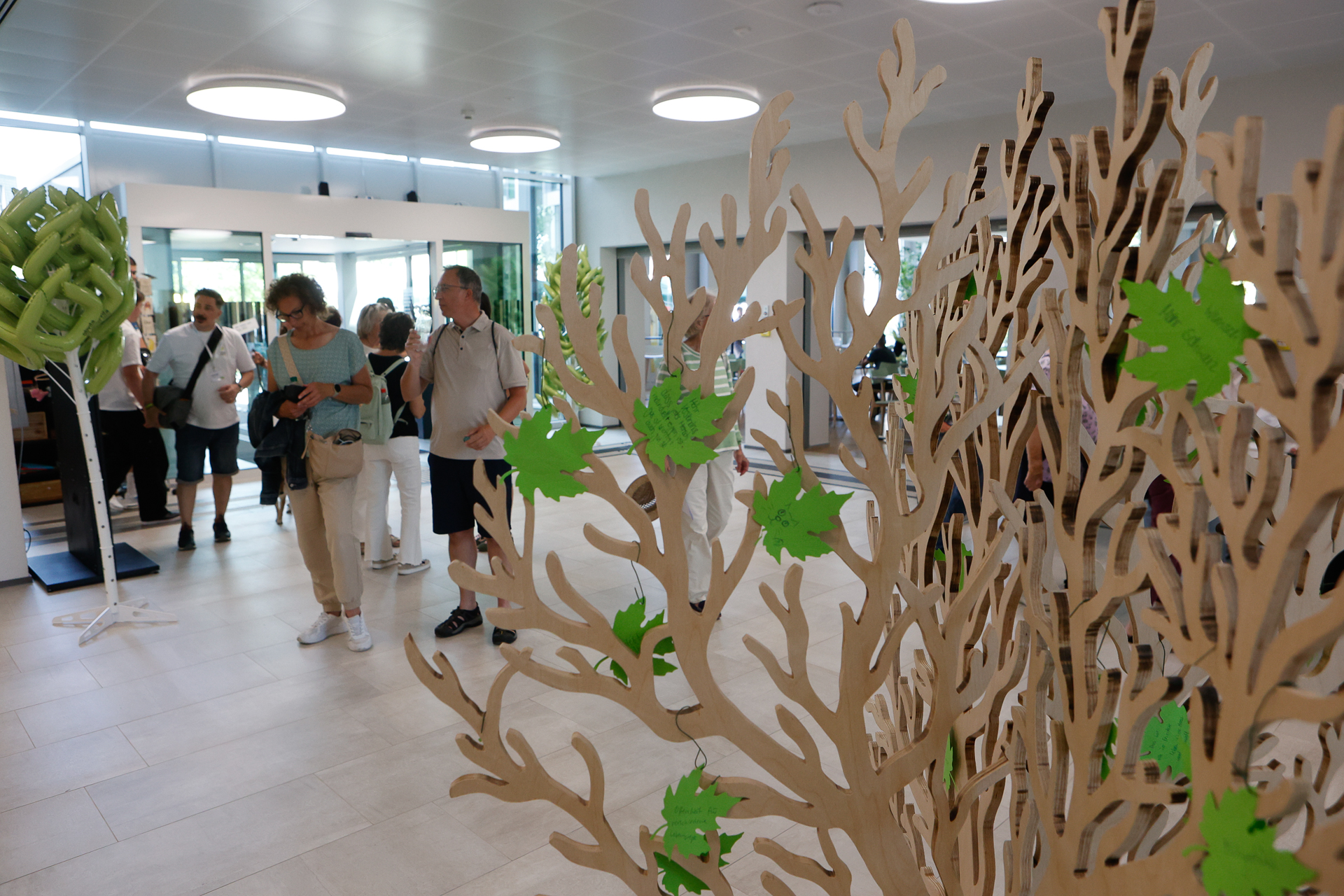 Im Vordergrund sind Menschen zu sehen, die durch einen hellen Innenraum gehen, in dem baumartige Holzskulpturen stehen, die mit grünen Papierblättern geschmückt sind. Der Raum verfügt über große Fenster und eine moderne Beleuchtung.