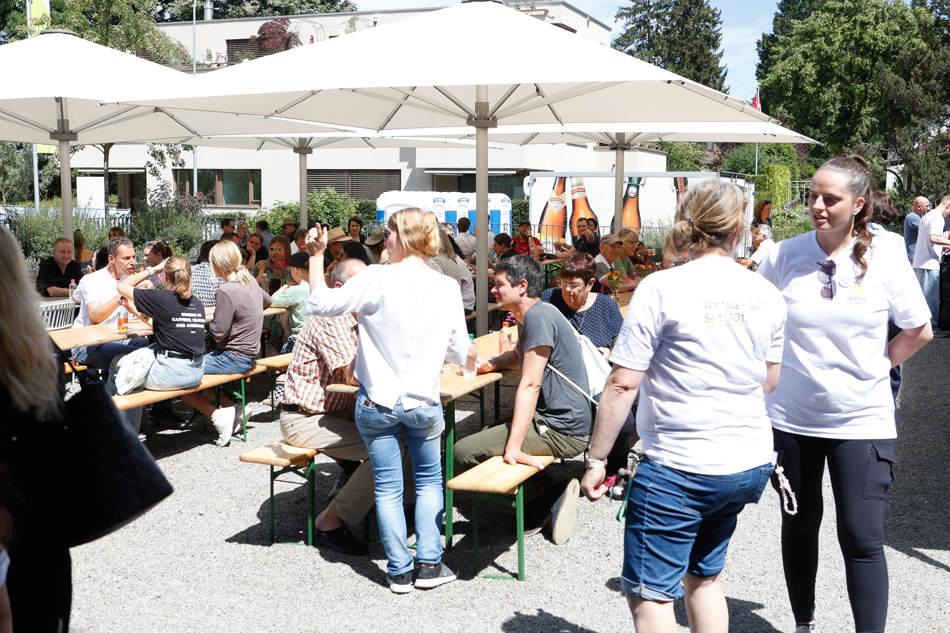 Eine Gruppe von Menschen sitzt an hölzernen Picknicktischen unter großen weißen Sonnenschirmen und genießt ein sonniges Beisammensein im Freien. Zwei Frauen in weißen T-Shirts gehen in der Nähe vorbei, während sich andere im Hintergrund unterhalten und entspannen.