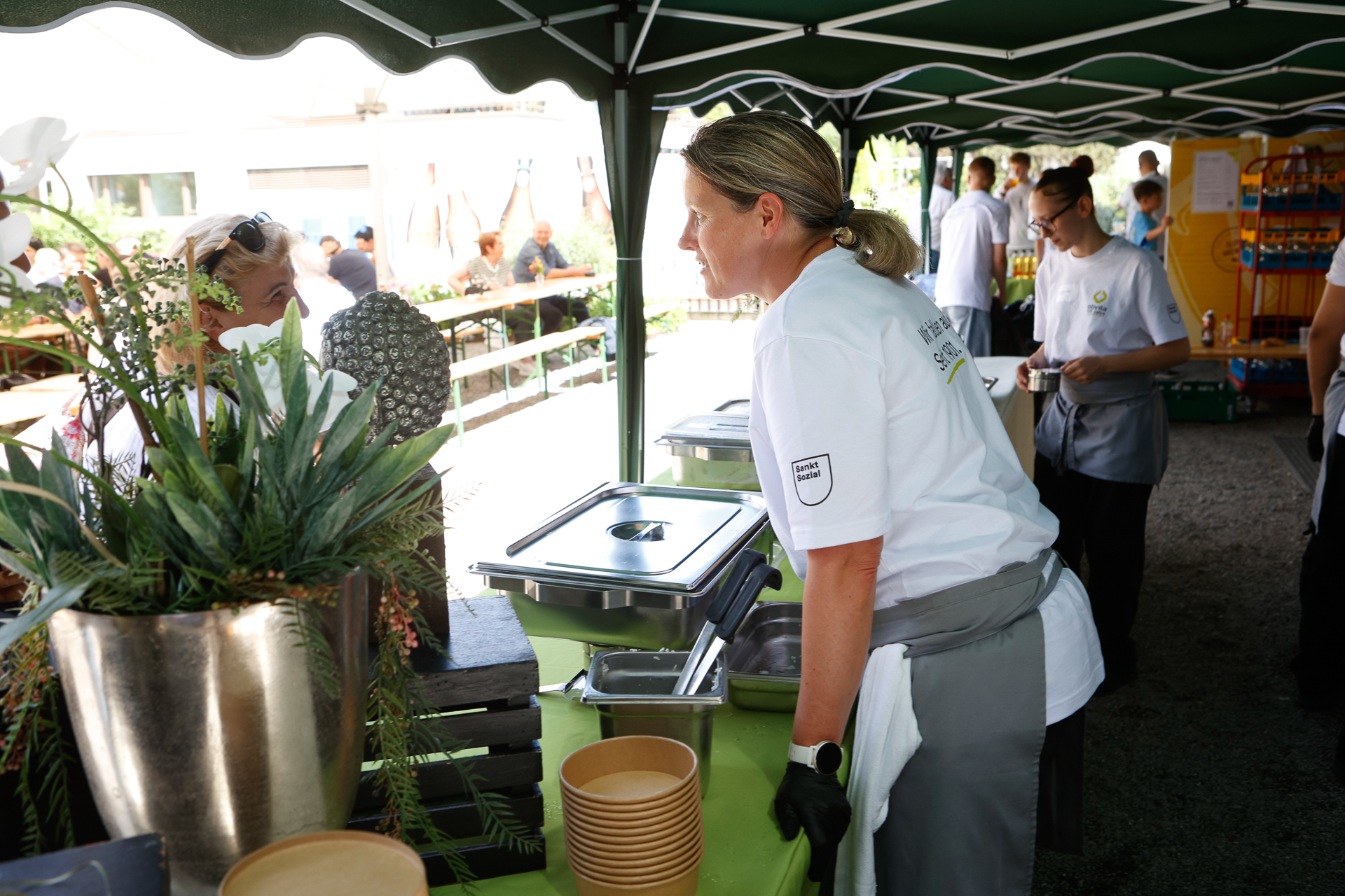 Eine Frau in einem weißen Hemd und einer grauen Schürze serviert Speisen an einem Buffet im Freien unter einem Vordach; im Hintergrund sind weitere Mitarbeiter und Gäste zu sehen. Im Vordergrund steht eine große Topfpflanze.