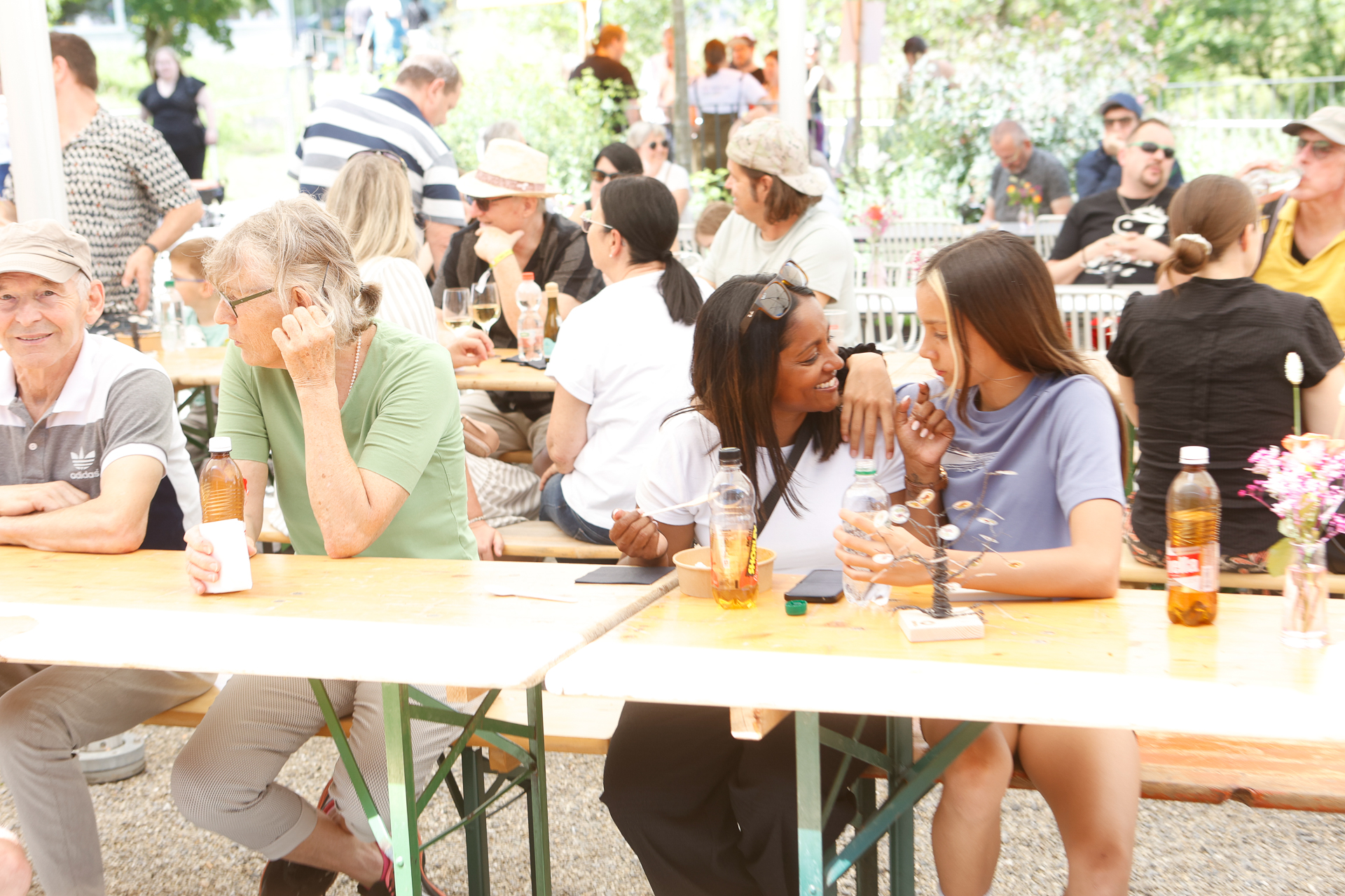 Die Leute sitzen draußen an hölzernen Picknicktischen, unterhalten sich und lächeln. In der Mitte lachen zwei Frauen gemeinsam, auf dem Tisch stehen Flaschen und Gläser. Im Hintergrund entspannen sich andere Gruppen unter einem schattigen Vordach und unterhalten sich.