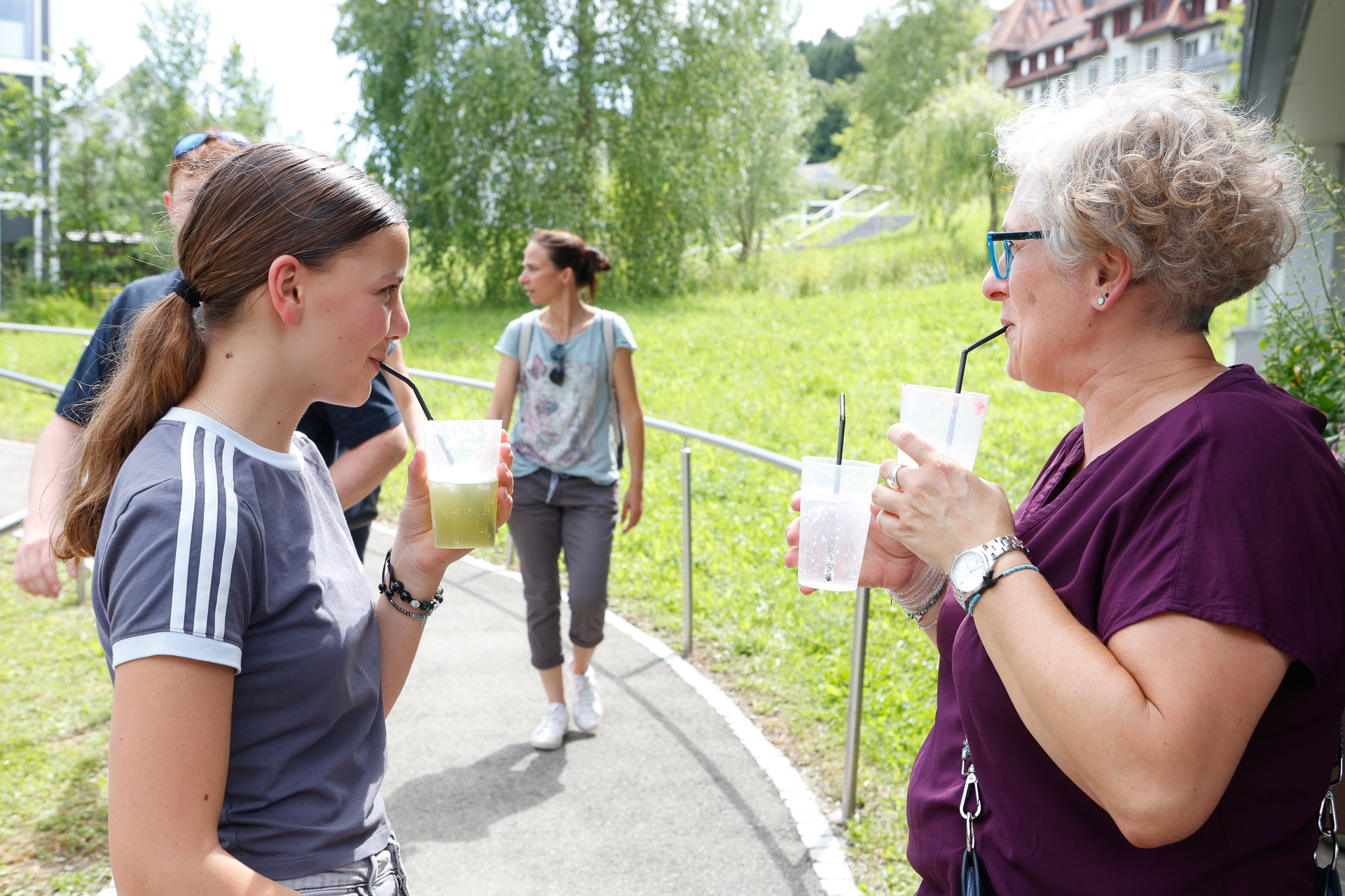 Zwei Frauen stehen an einem sonnigen Tag im Freien, trinken aus Plastikbechern mit Strohhalmen und unterhalten sich. Im Hintergrund sind andere Menschen zu sehen, die an grünem Gras und Bäumen vorbeigehen. Eine Frau trägt eine Sonnenbrille und ein lila Shirt, die andere ein grau gestreiftes Shirt.
