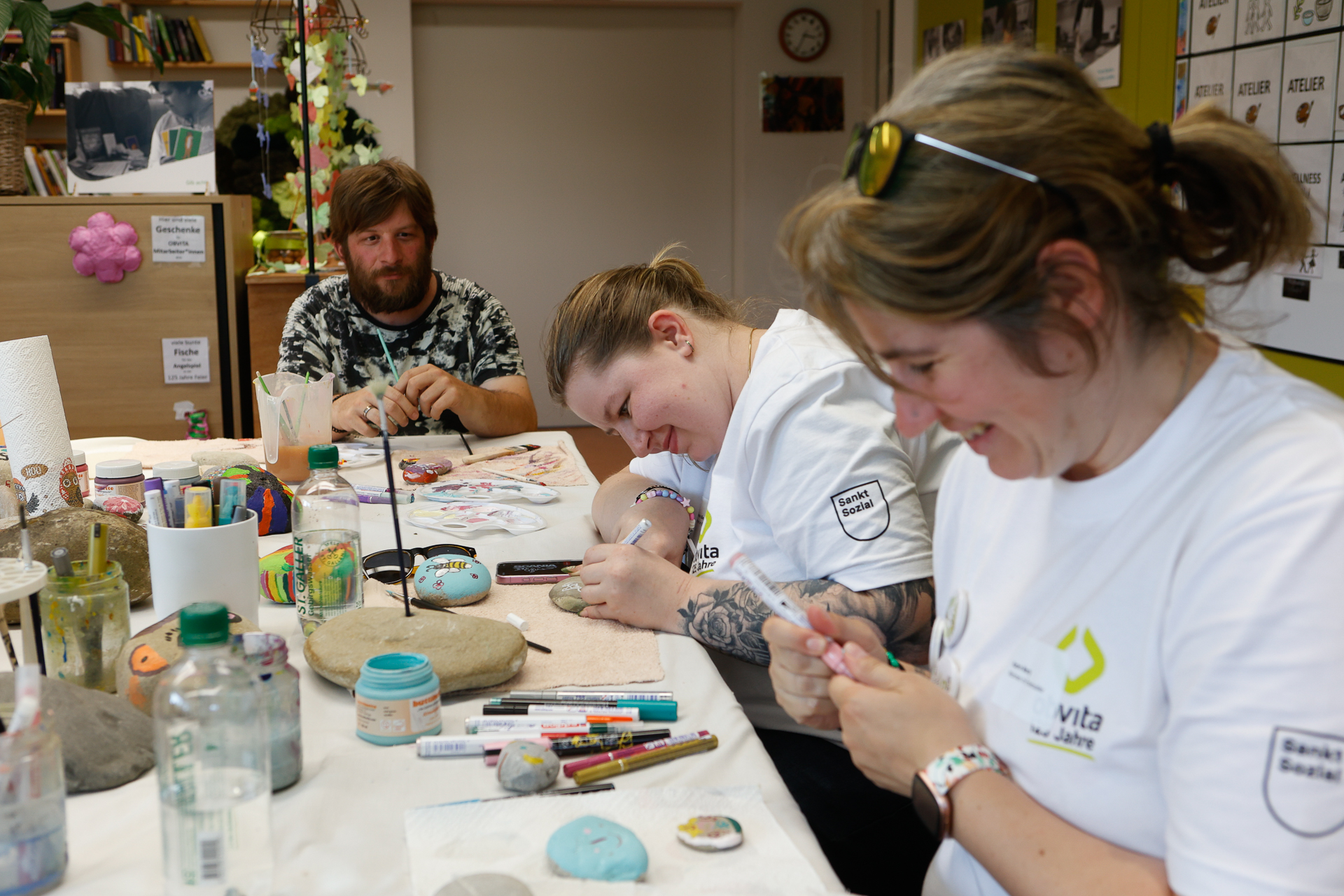 Drei Menschen sitzen an einem Tisch, der mit Malutensilien bedeckt ist, bemalen Steine und lachen gemeinsam. Zwei Frauen in weißen Hemden konzentrieren sich auf ihre Arbeit, während ein Mann in einem gemusterten Hemd im Hintergrund zusieht und lächelt.