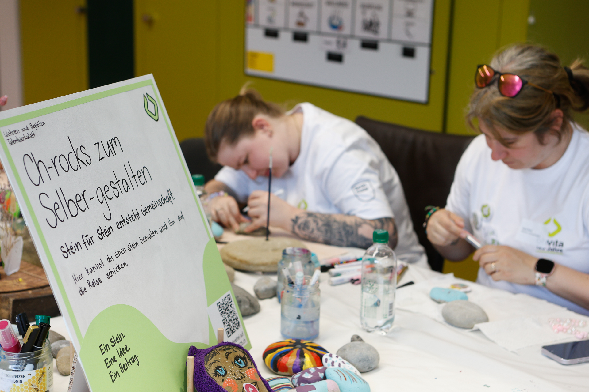 Zwei Personen sitzen an einem Tisch und verzieren Steine mit bunten Motiven. Im Vordergrund lädt ein Schild auf Deutsch die Teilnehmer dazu ein, ihre eigenen Steine zu bemalen. Auf dem Tisch liegen Malutensilien und fertig bemalte Steine.