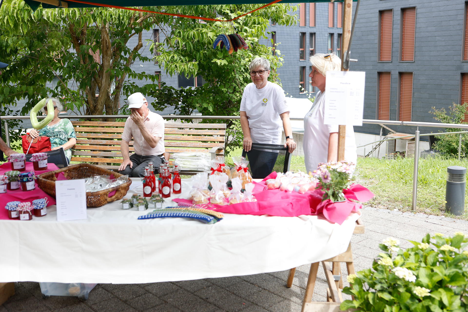 Ein Marktstand im Freien, auf dem Gläser mit Marmelade, Flaschen und Kekse ausgestellt sind. Zwei Personen stehen hinter dem Tisch, eine Person sitzt auf einer Bank. An einem Ende des Tisches sind rosa Blumen und Geschenkartikel arrangiert.