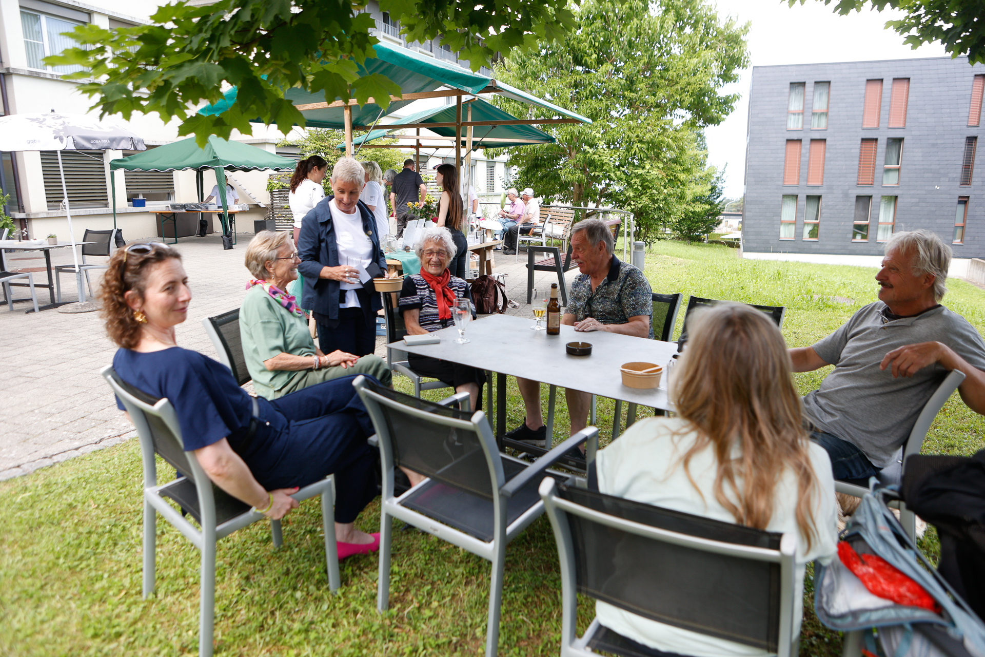 Eine Gruppe von Menschen sitzt und steht im Freien auf einer Wiese um einen Tisch herum, unterhält sich und lächelt. Im Hintergrund sind Bäume, Sonnenschirme und moderne Gebäude zu sehen. Einige der Menschen halten Getränke in der Hand.