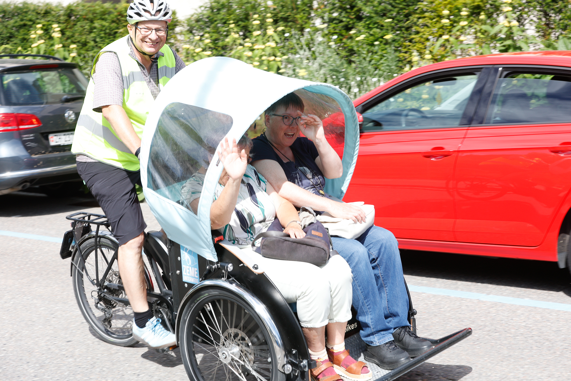 Ein Mann mit Helm und Warnweste tritt in die Pedale einer blauen Fahrradrikscha, auf der zwei lächelnde ältere Frauen unter einem Sonnendach sitzen; eine der Frauen winkt, während Autos auf der Straße vorbeifahren.