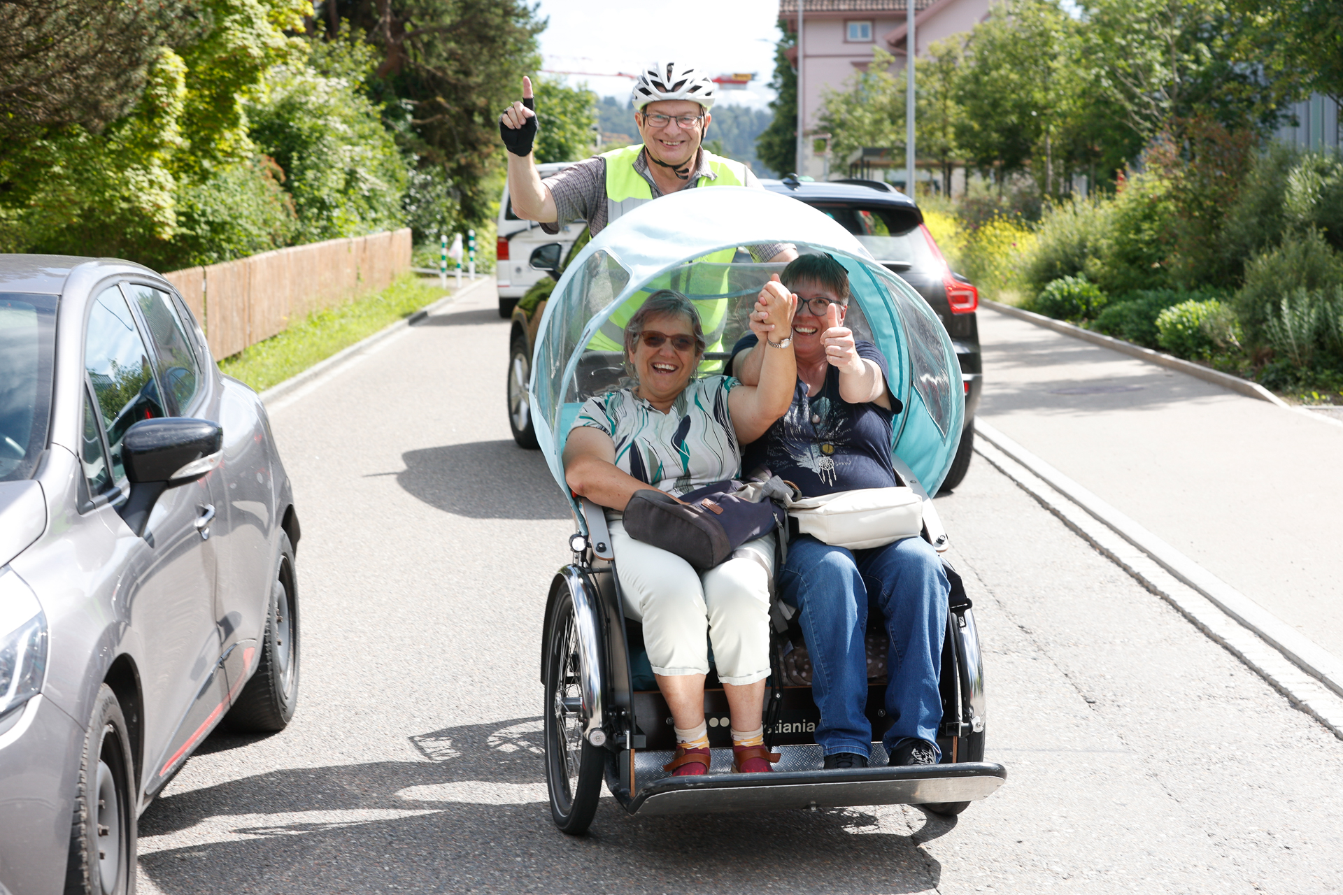 Zwei Frauen sitzen auf dem Vordersitz eines Dreirads mit durchsichtiger Abdeckung, lächeln und zeigen den Daumen nach oben, während ein Mann hinter ihnen auf einer sonnigen Straße mit Autos und viel Grün in der Umgebung in die Pedale tritt.