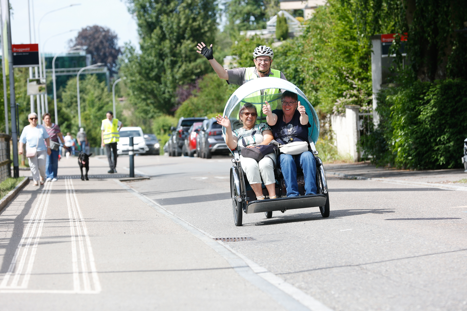 Ein Mann mit Helm und Fahrradbekleidung tritt in eine Rikscha mit Verdeck, in der zwei lächelnde Passagiere sitzen, die winken, während sie auf einer sonnigen Stadtstraße fahren. Auf dem Bürgersteig gehen Menschen spazieren, und entlang der Straße stehen Autos geparkt.