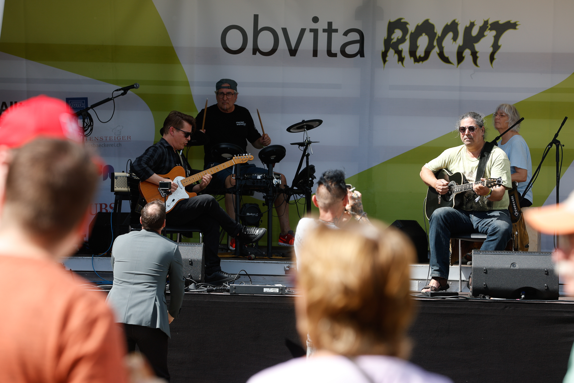 Eine Band tritt auf einer Open-Air-Bühne mit E-Gitarren, Akustikgitarren und Schlagzeug auf, während das Publikum zusieht. Im Hintergrund ist in fetten Buchstaben der Schriftzug „obvita ROCKT“ zu sehen.