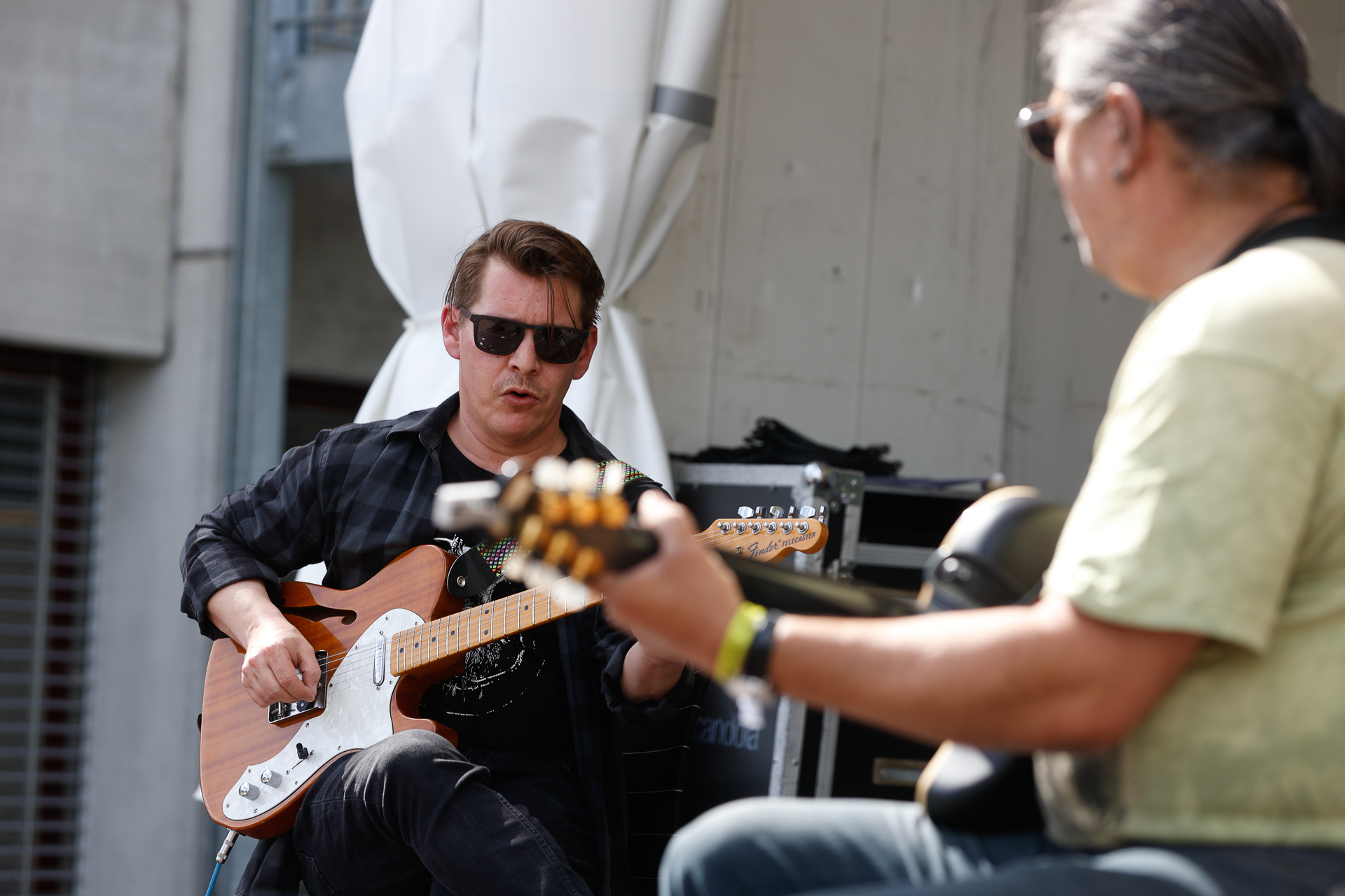 Zwei Männer sitzen im Freien und spielen E-Gitarre. Ein Mann mit Sonnenbrille und kariertem Hemd konzentriert sich auf sein Instrument, während der andere, mit langen Haaren und einem grünen Hemd, neben ihm klimpern lässt. Hinter ihnen sind Verstärker und Equipment zu sehen.