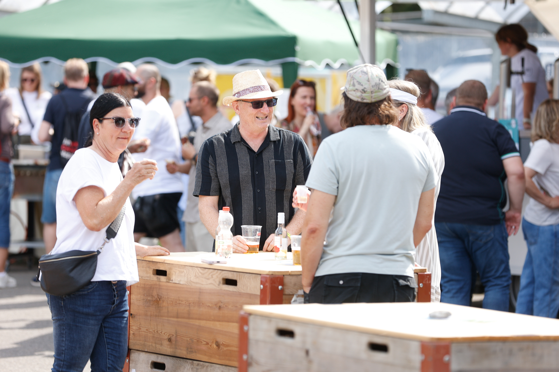 Eine Gruppe von Menschen, die sich im Freien auf einem Markt oder einem Fest unterhält und mit Getränken um Holztische herumsteht. Einige tragen Sonnenbrillen und Hüte, und im Hintergrund ist ein grünes Zeltdach zu sehen.
