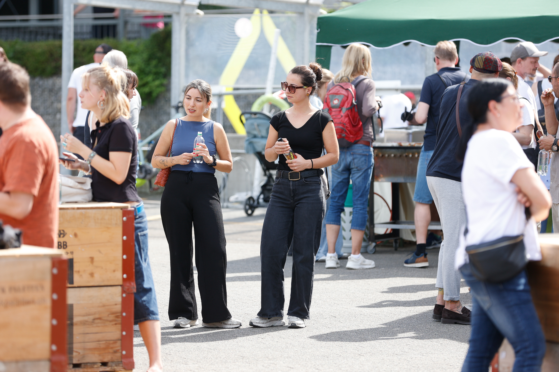 Bei einer Veranstaltung versammeln sich die Menschen im Freien. Zwei Frauen in schwarzen Oberteilen und Hosen stehen nebeneinander und halten Getränke in der Hand. Andere Teilnehmer unterhalten sich in der Nähe unter einem grünen Vordach. Die Atmosphäre ist ungezwungen und sonnig.