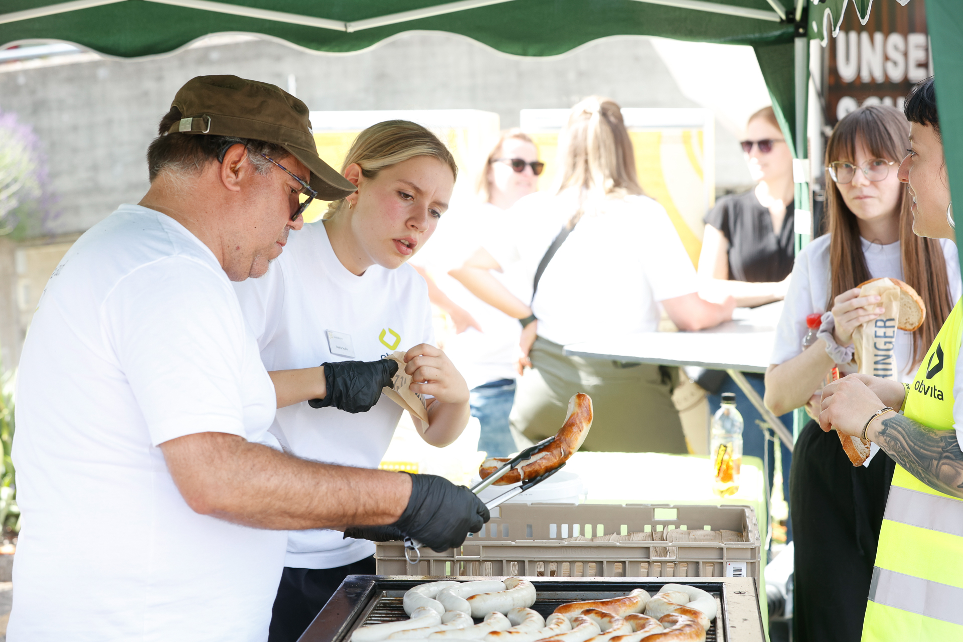 Bei einer Veranstaltung im Freien grillen zwei Personen unter einem grünen Vordach Brezeln und Hot Dogs, während andere in weißen Hemden im Hintergrund an einem Tisch stehen und sitzen.