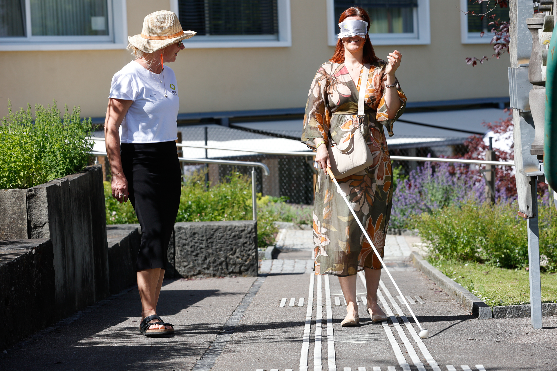 Eine Frau in einem gemusterten Kleid hat die Augen verbunden und bewegt sich mit einem Blindenstock auf einem taktilen Pfad entlang, während eine andere Frau mit Hut und weißem Hemd sie im Freien beobachtet.