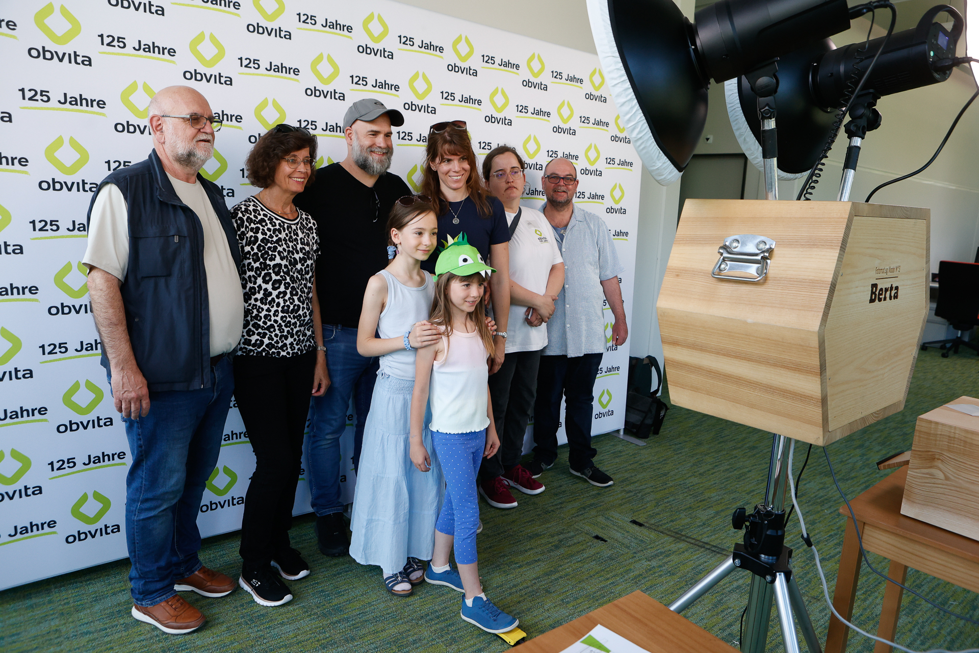 Eine Gruppe aus sieben Erwachsenen und zwei Kindern posiert für ein Foto vor einem Hintergrund mit der Aufschrift „125 Jahre obvita“. Im Vordergrund sind eine professionelle Beleuchtung und eine Kamera aufgebaut.