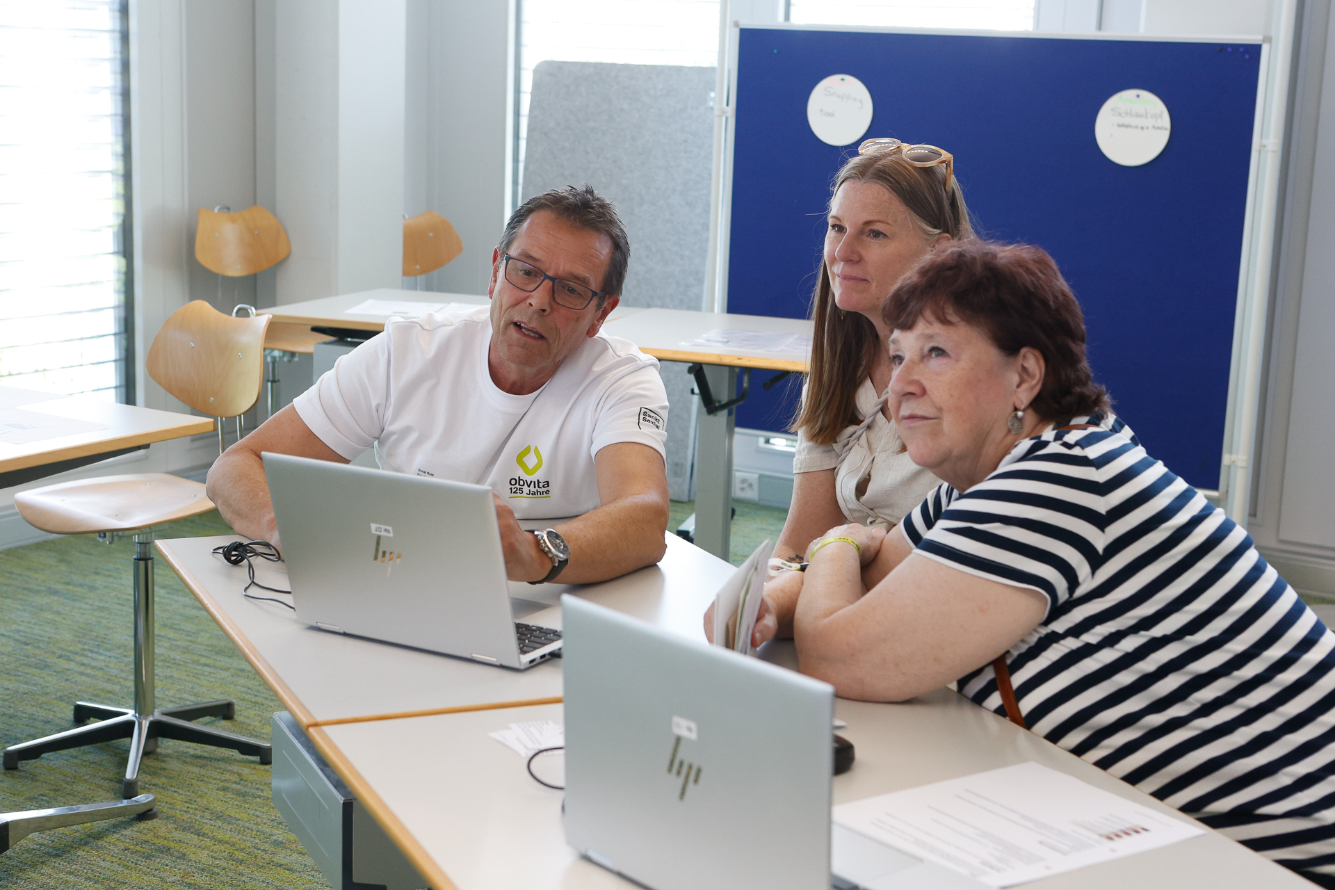 Drei Erwachsene sitzen gemeinsam an einem Schreibtisch mit Laptops und scheinen gemeinsam zu arbeiten. Zwei Frauen und ein Mann blicken konzentriert auf einen Bildschirm in einem hellen, modernen Klassenzimmer.