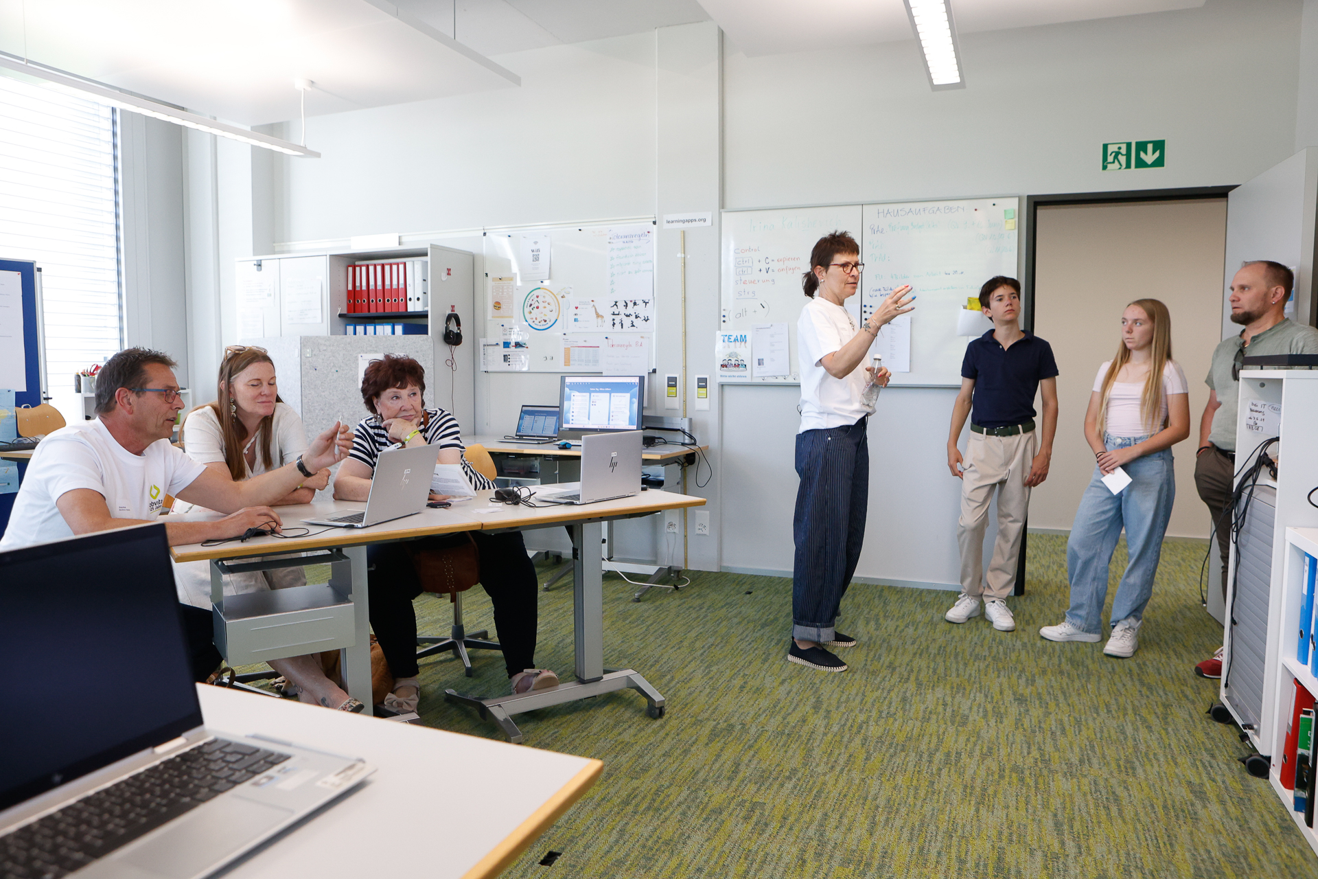 Eine Gruppe von Menschen arbeitet an Laptops um einen Tisch herum, während eine andere Gruppe neben einem Whiteboard steht. Eine Person spricht zu der stehenden Gruppe in einem modernen, hell beleuchteten Büro oder Klassenzimmer.