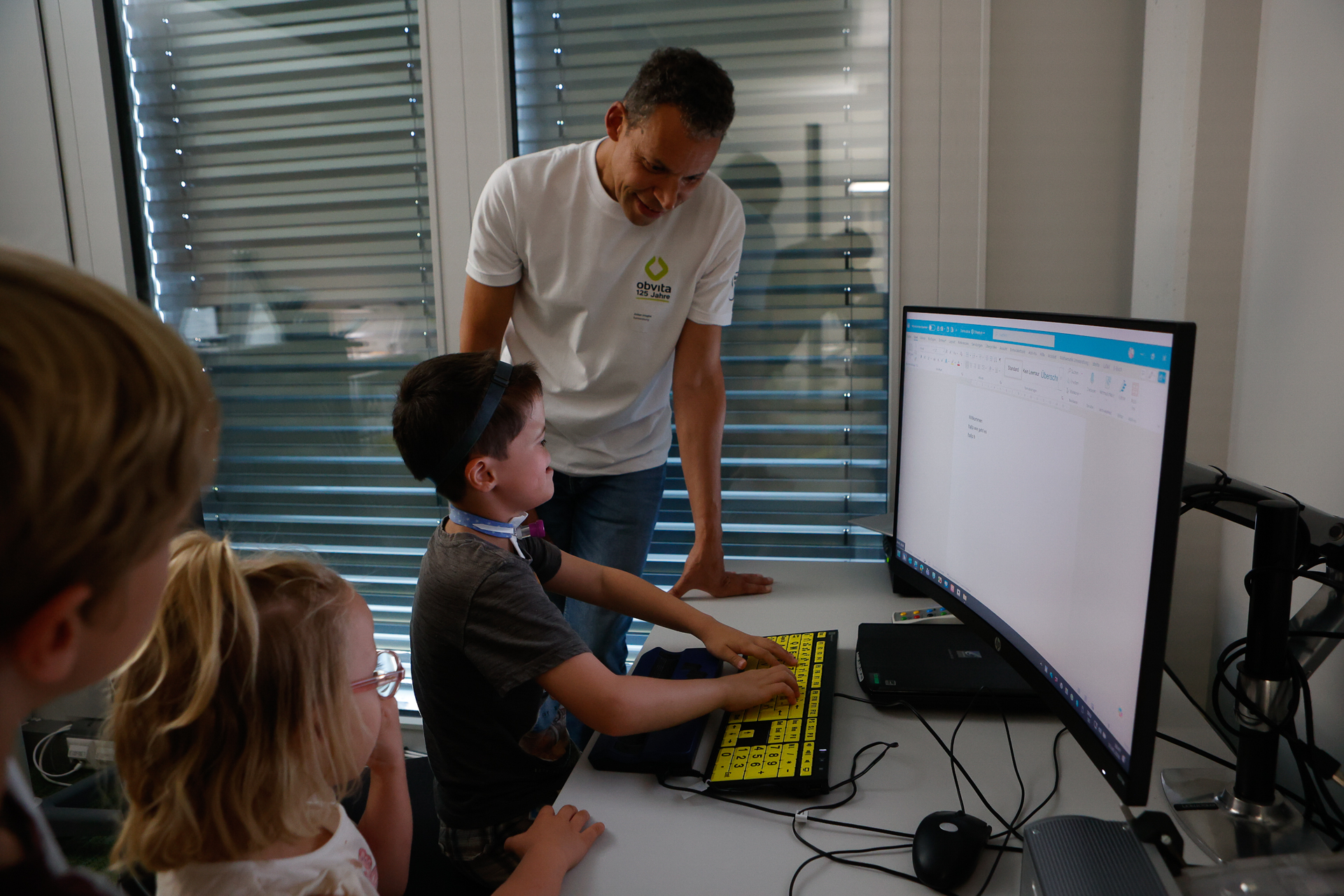 Ein Mann steht neben einem Jungen, der an einer großen gelben Tastatur und einem Computer sitzt, während zwei weitere Kinder zuschauen. Sie befinden sich in einem hellen Raum, dessen Fenster mit Jalousien verdeckt sind.