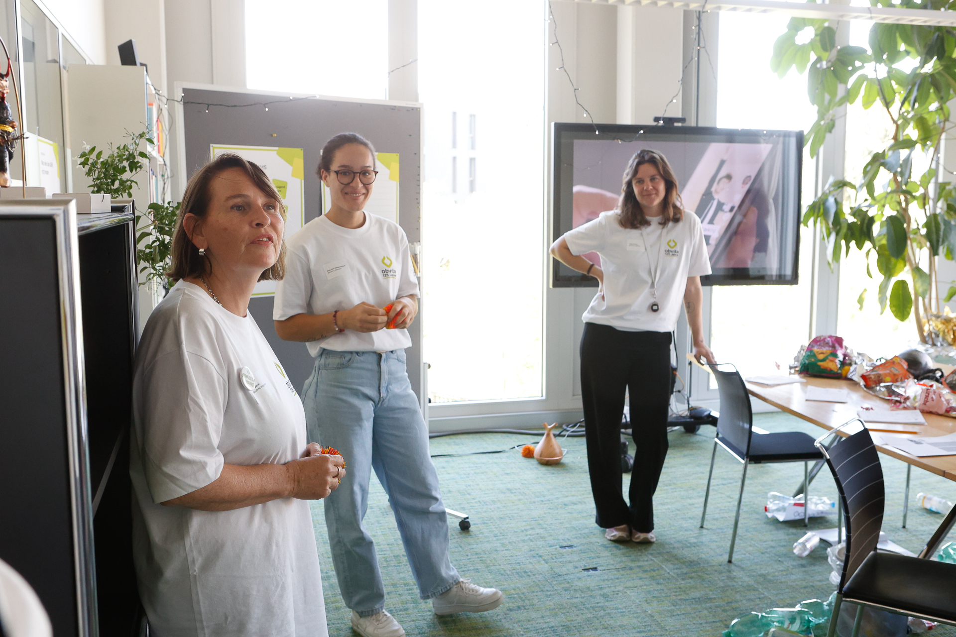 Drei Frauen in weißen T-Shirts stehen in einem hellen, modernen Raum mit großen Fenstern. Eine spricht, während die anderen zuhören und lächeln. Im Hintergrund stehen Stühle, Pflanzen und ein Paravent, und überall liegen verschiedene Gegenstände verstreut.