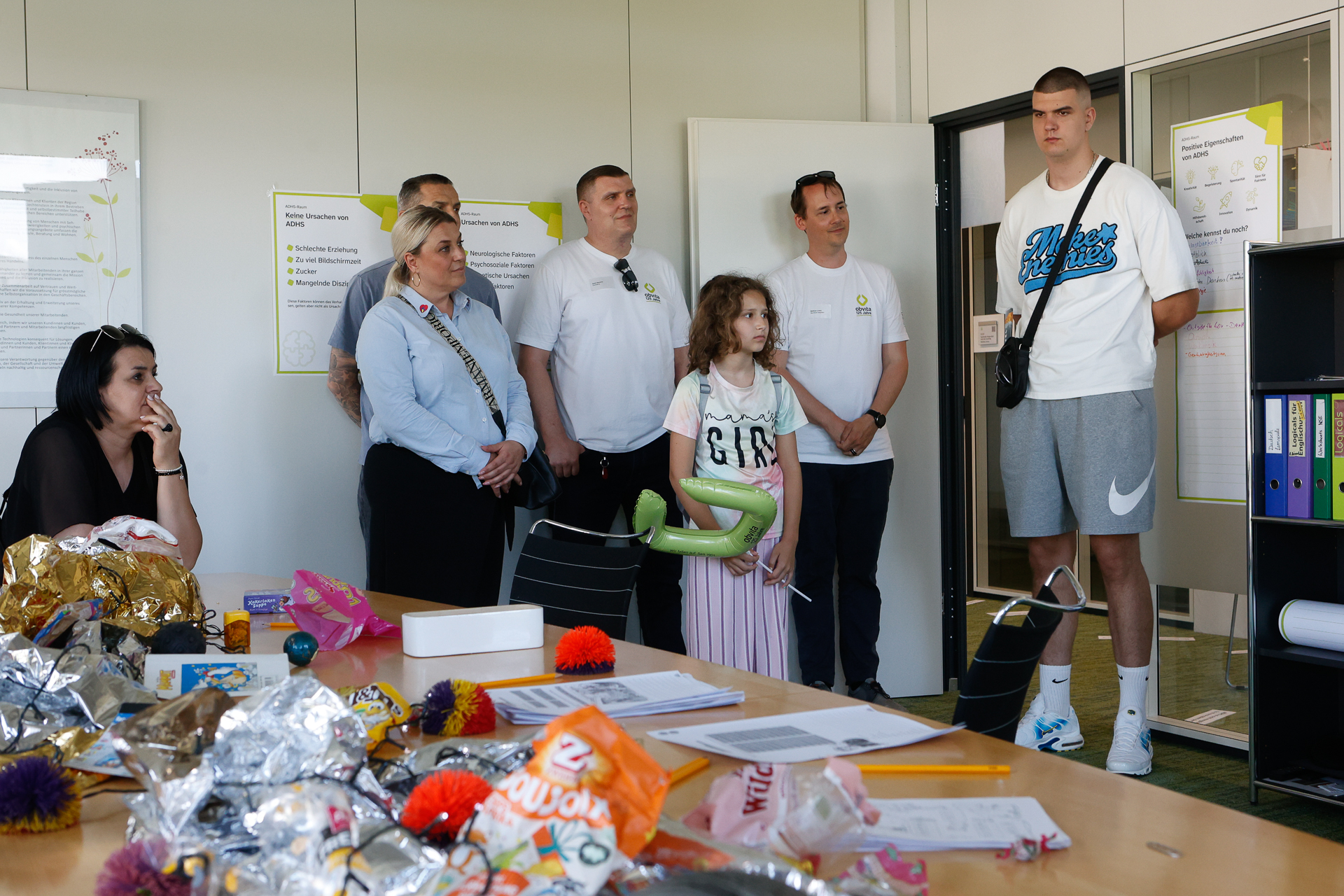 Eine Gruppe von Erwachsenen und ein Kind stehen in einem Raum neben einem Tisch, auf dem Bastelmaterial und Papier liegen, während eine Frau in der Nähe sitzt. Die Stimmung wirkt aufmerksam, als würden alle jemandem zuhören.