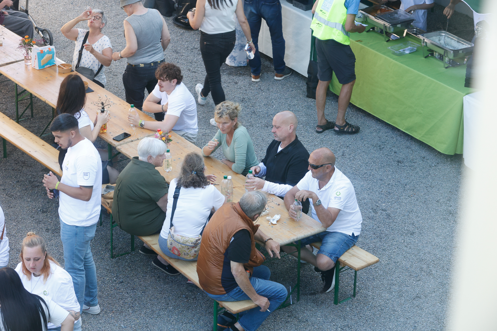 Eine Gruppe von Menschen sitzt und steht im Freien um hölzerne Picknicktische herum, isst, trinkt und unterhält sich. Das Essen wird von einem grün gedeckten Tisch in der Nähe serviert. Einige tragen weiße Hemden und Westen. Die Atmosphäre wirkt ungezwungen und gesellig.