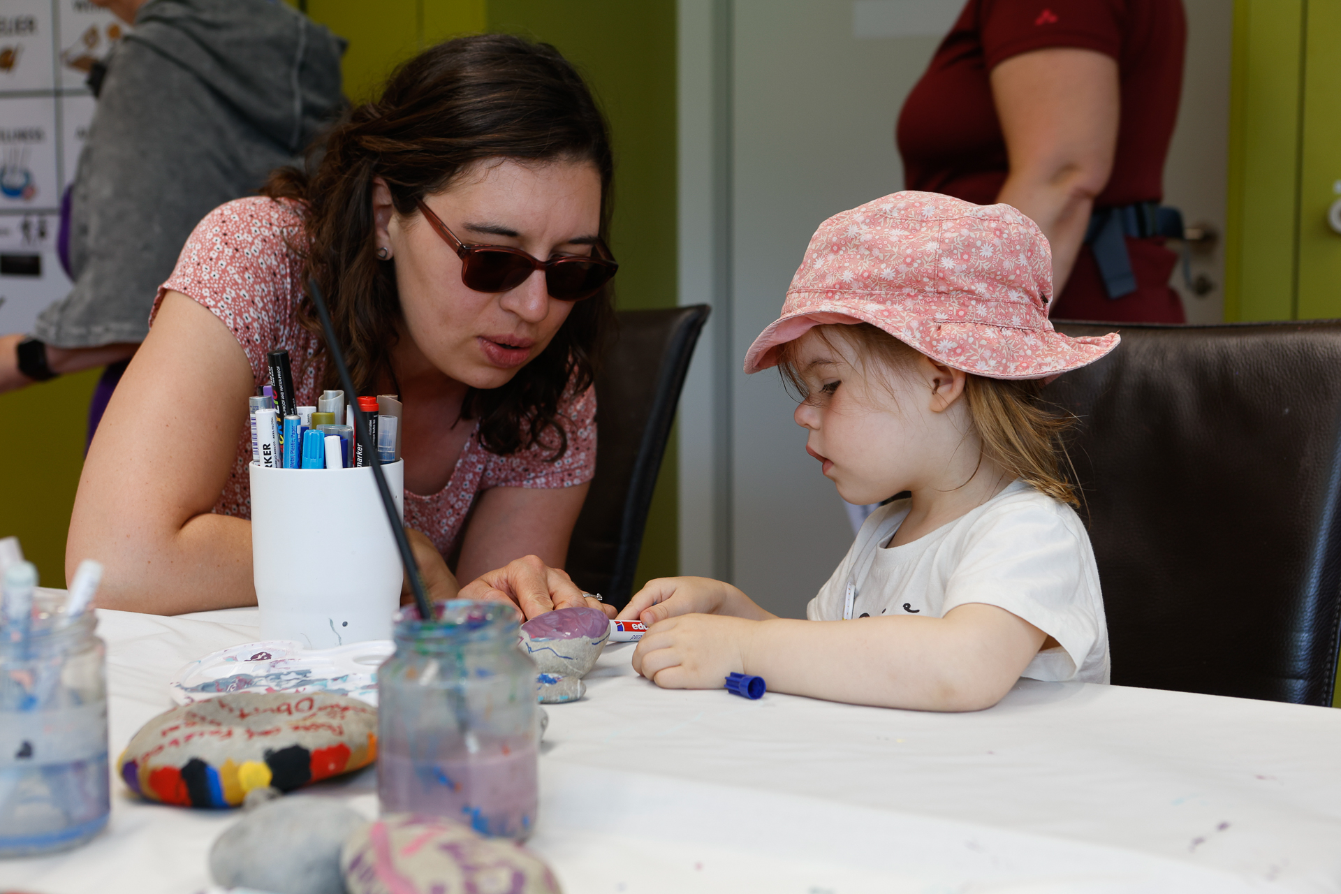 Eine Frau mit Sonnenbrille hilft einem kleinen Mädchen mit rosa Mütze bei einer Bastelarbeit an einem Tisch, auf dem bemalte Steine, Filzstifte und Gläser liegen. Beide sind ganz in ihre Arbeit vertieft.