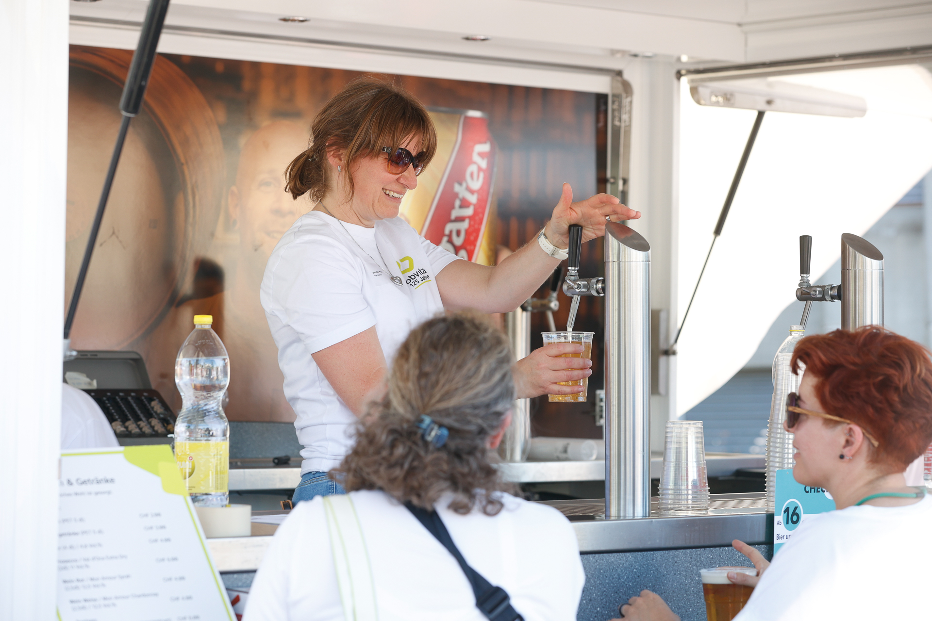 Eine lächelnde Frau mit Sonnenbrille serviert drei Frauen an einem Getränkestand im Freien ein Getränk. Sie hält einen Plastikbecher mit Bier in der Hand, während sie sich mit den Kundinnen unterhält. Auf der Theke sind eine Speisekarte und Flaschen zu sehen.