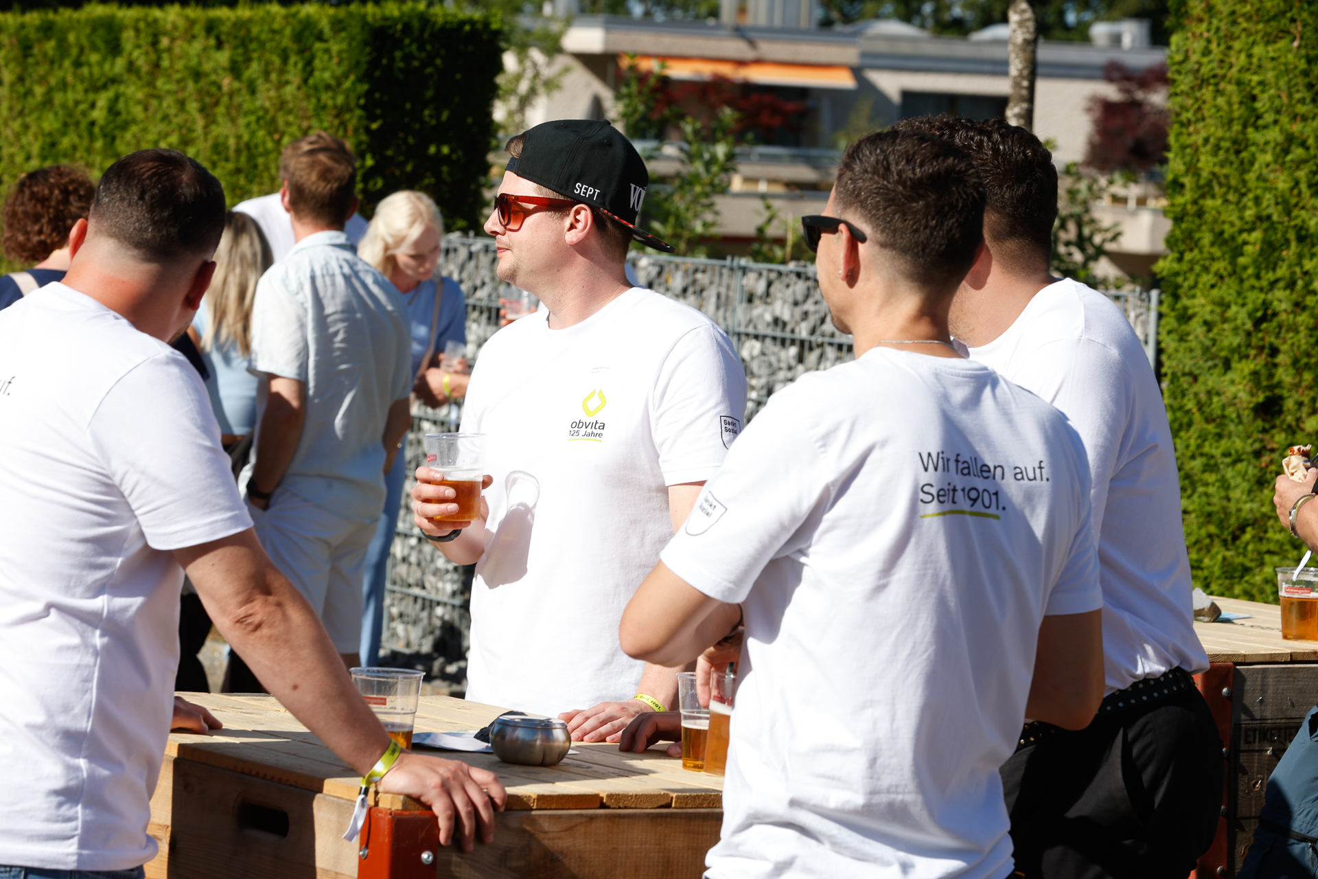 Eine Gruppe von Männern in weißen Hemden steht im Freien um einen Holztisch herum, hält Bierkrüge in den Händen und unterhält sich, während sich im Hintergrund an diesem sonnigen Tag andere unterhalten.
