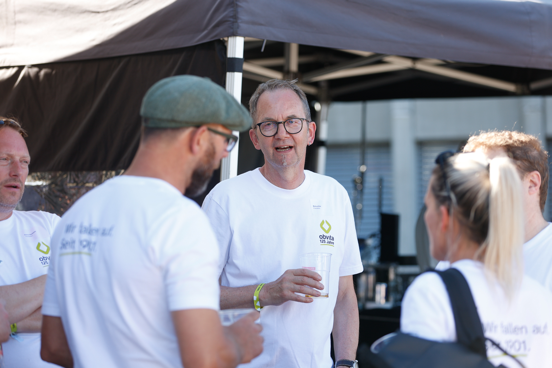Eine Gruppe von Menschen in weißen Hemden steht draußen beieinander, unterhält sich und hält Getränke in den Händen. Ein Mann in der Mitte spricht, während die anderen zuhören. Sie befinden sich an einem sonnigen Tag unter einem schwarzen Festzelt.