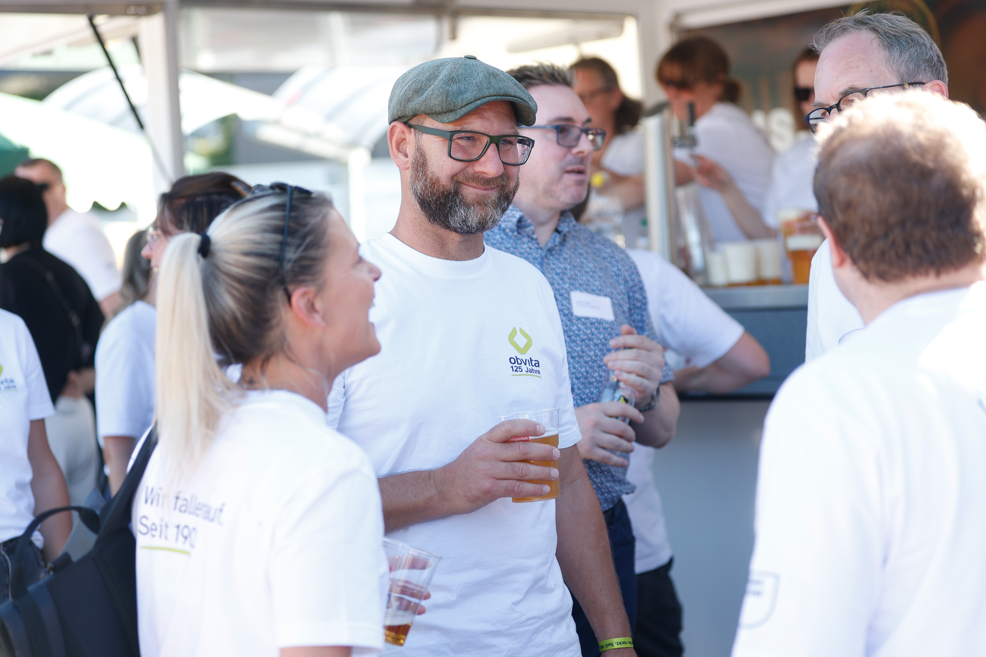 Eine Gruppe von Menschen in Freizeitkleidung unterhält sich bei einer Veranstaltung im Freien und hält Getränke in der Hand; ein Mann mit grüner Mütze und Brille lächelt eine Frau im Vordergrund an.