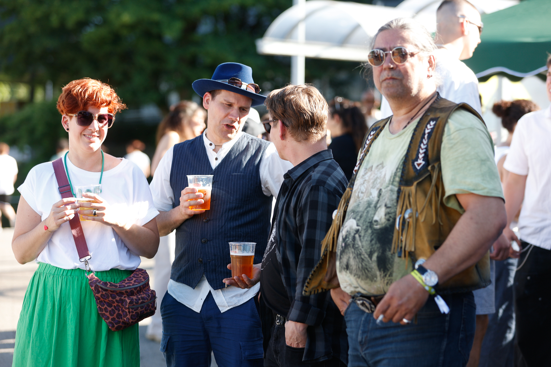 Eine Gruppe von Menschen steht bei einer Veranstaltung im Freien in der Sonne, hält Getränke in der Hand und unterhält sich. Einige tragen Freizeitkleidung, während andere ausgefallene Accessoires wie Hüte und Westen tragen. Im Hintergrund sind Bäume und weitere Menschen zu sehen.