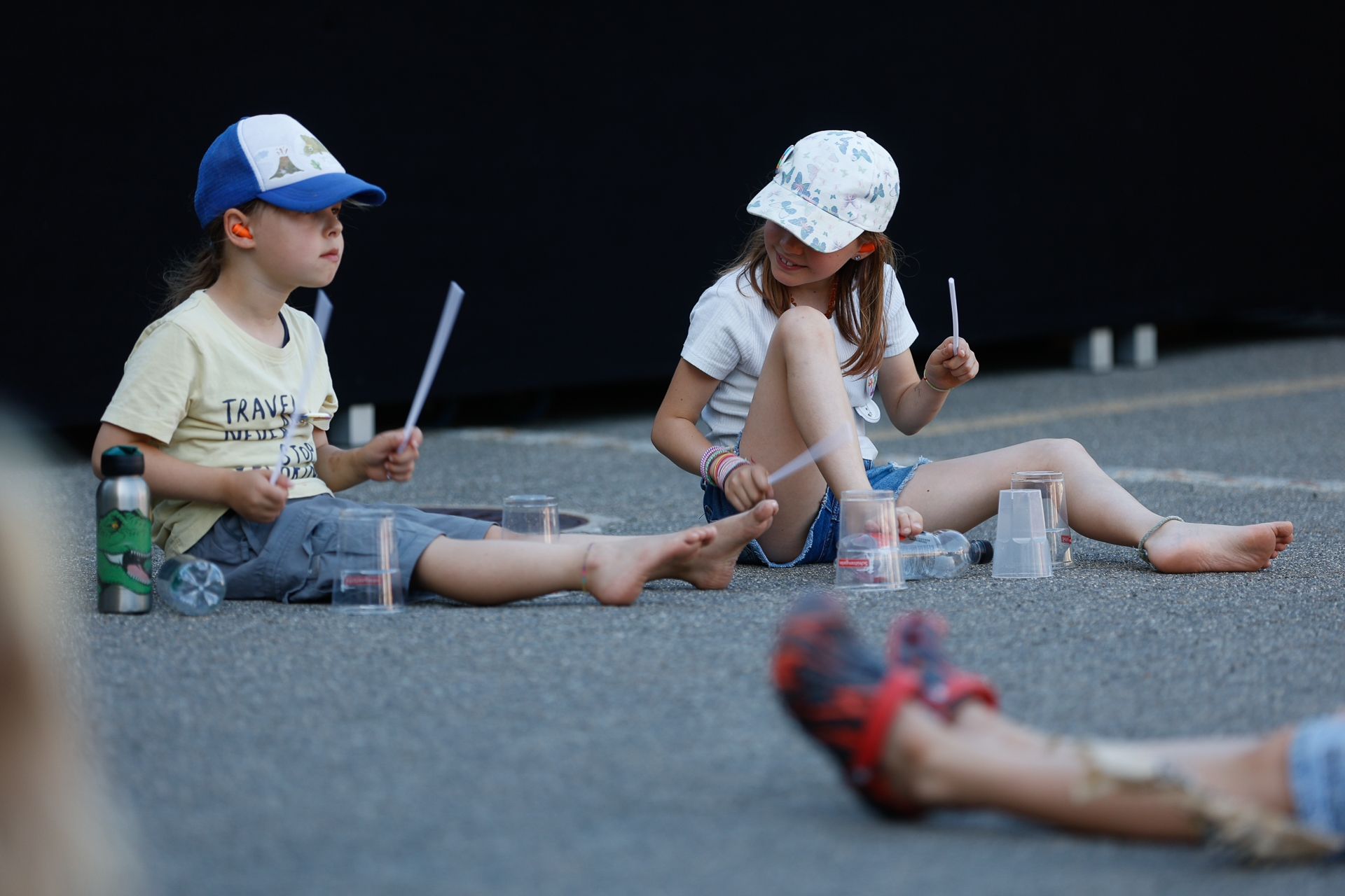 Zwei Kinder mit Mützen sitzen auf dem Boden und machen mit Plastikbechern und Stöcken Musik. Sie sind barfuß, neben ihnen stehen Wasserflaschen, und sie sind auf einer gepflasterten Fläche mit einer spielerischen Aktivität beschäftigt.