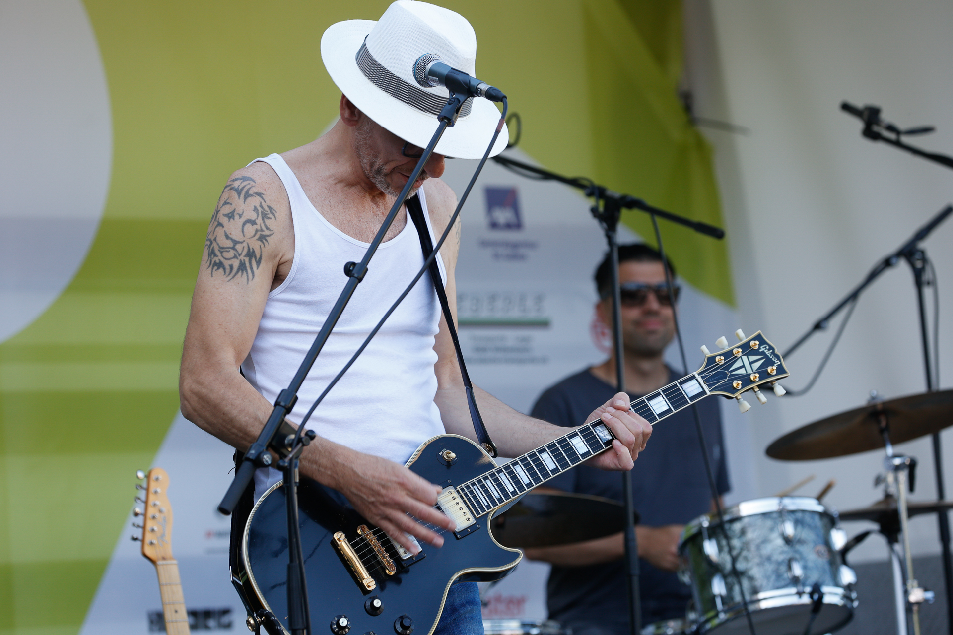 Ein Musiker mit weißem Hut und Tanktop spielt auf der Bühne E-Gitarre, im Hintergrund ist ein Schlagzeuger zu sehen. Mikrofone und Musikausrüstung sind zu erkennen, und hinter ihnen befindet sich ein grün-weißer Hintergrund.
