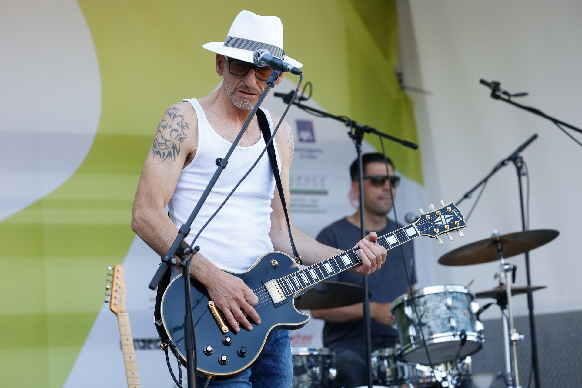 Ein Musiker, der einen weißen Hut, eine Sonnenbrille und ein weißes Tanktop trägt, spielt auf der Bühne E-Gitarre und singt ins Mikrofon, während im Hintergrund ein Schlagzeuger spielt.