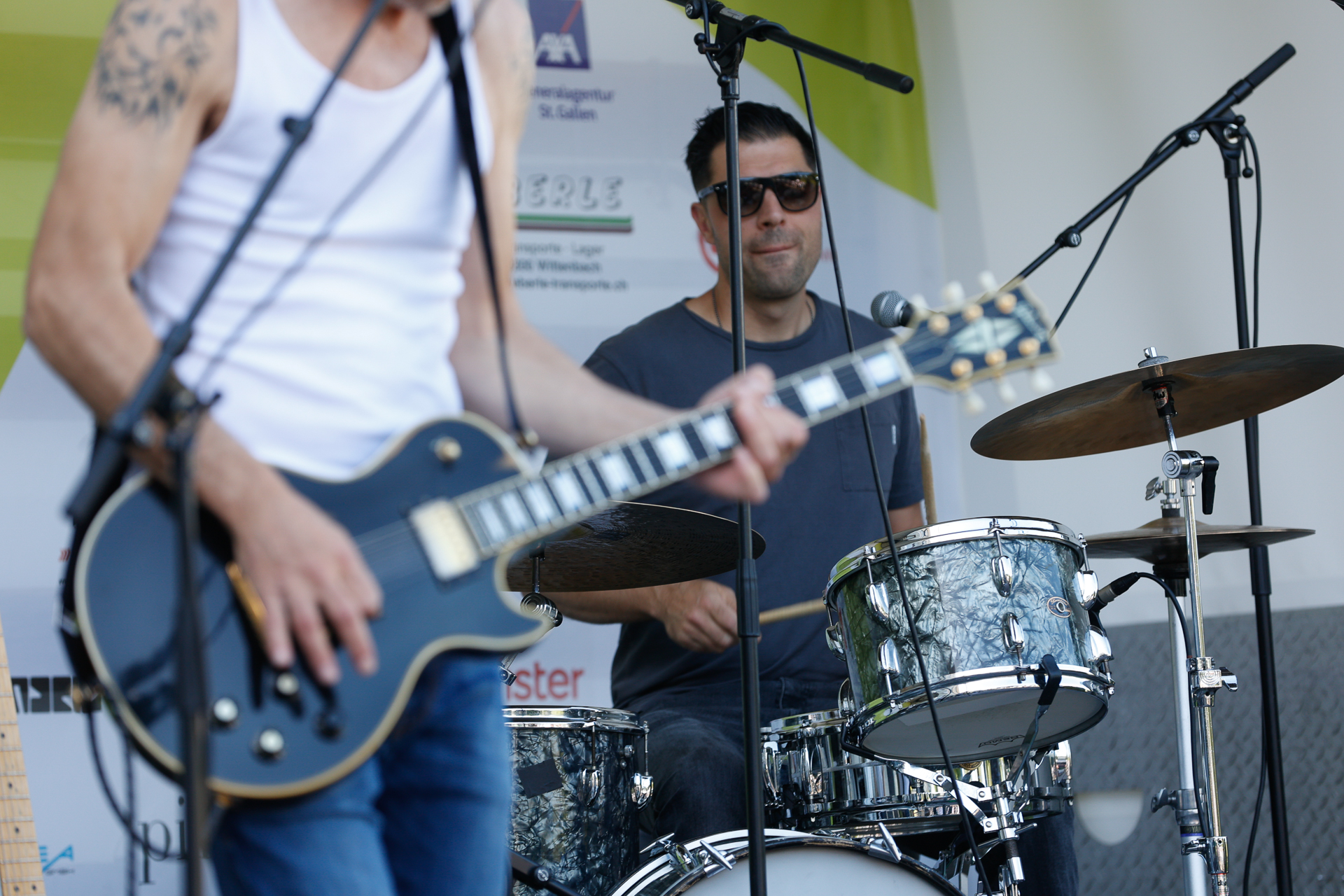 Ein Musiker in einem weißen Tanktop spielt E-Gitarre, während eine andere Person mit Sonnenbrille und dunklem Hemd auf einer Außenbühne Schlagzeug spielt. Mikrofone und Musikinstrumente sind zu sehen.