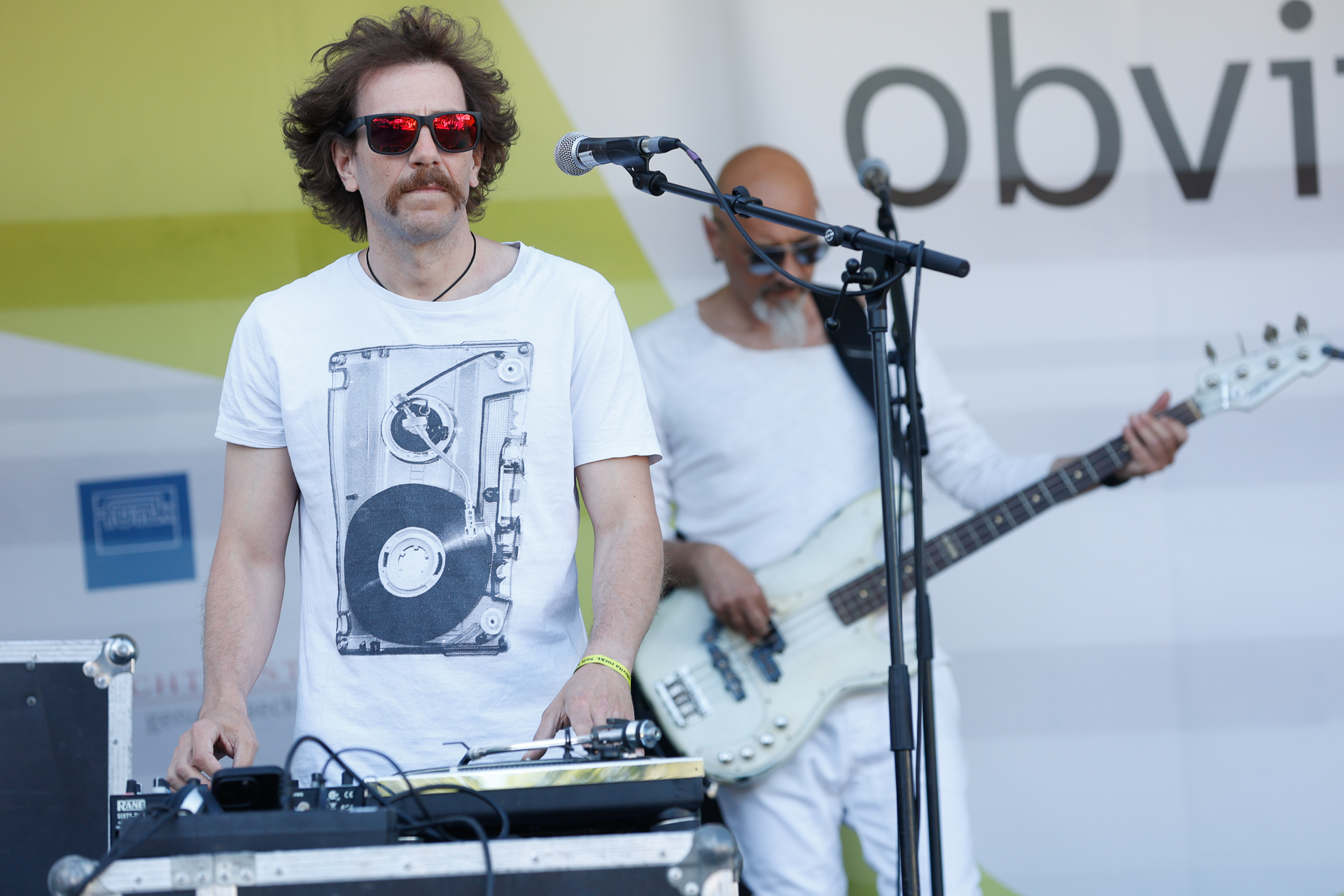 Zwei Musiker treten auf der Bühne auf. Der Musiker im Vordergrund, mit lockigem Haar und Sonnenbrille, bedient die DJ-Anlage und trägt ein weißes T-Shirt mit einem Plattenspieler-Motiv. Der zweite Musiker in Weiß spielt im Hintergrund Bassgitarre.