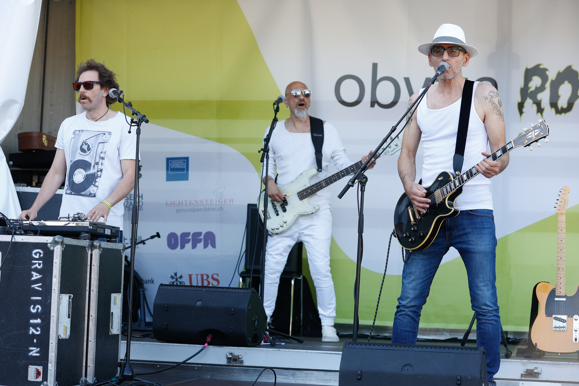 Drei männliche Musiker treten auf einer Freiluftbühne auf. Einer singt und spielt dabei Gitarre; er trägt einen weißen Hut und ein Tanktop. Ein anderer spielt Bass und ist ganz in Weiß gekleidet, und ein dritter spielt Keyboard. Hinter ihnen sind Sponsorenlogos zu sehen.