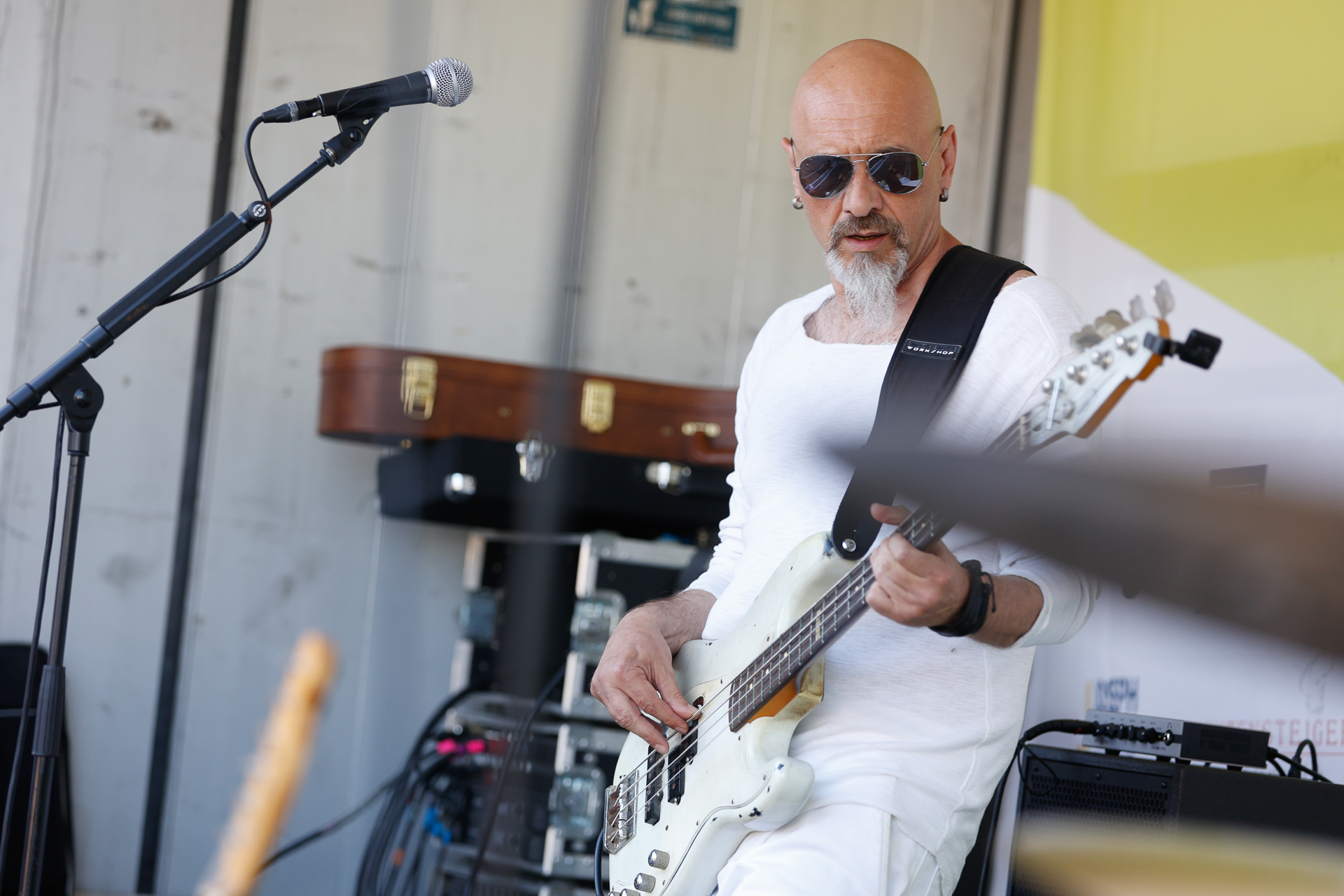 Ein kahlköpfiger Mann mit weißem Ziegenbart, der eine Sonnenbrille trägt und ganz in Weiß gekleidet ist, spielt auf einer Außenbühne in der Nähe eines Mikrofonständers auf einem weißen E-Bass. Im Hintergrund sind Musikinstrumente und ein Gitarrenkoffer zu sehen.