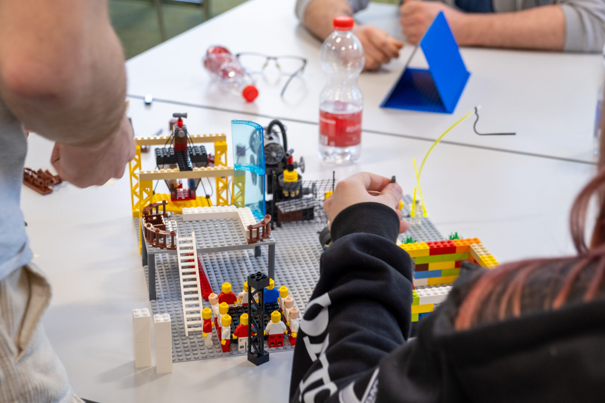 Auf einem Tisch wird ein buntes LEGO-Bauwerk errichtet. Mehrere LEGO-Figuren und -Steine sind angeordnet, und Hände nehmen Anpassungen an der Konstruktion vor. Im Hintergrund stehen Wasserflaschen, Gläser und Papiere.