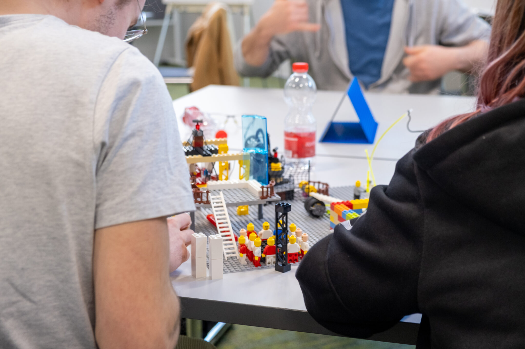 Zwei Personen sitzen an einem Tisch und bauen mit kleinen Figuren ein buntes LEGO-Modell. Ihnen gegenüber sitzt eine weitere Person, die im Hintergrund verschwommen zu sehen ist; auf dem Tisch stehen eine Wasserflasche und ein blaues, dreieckiges Objekt.