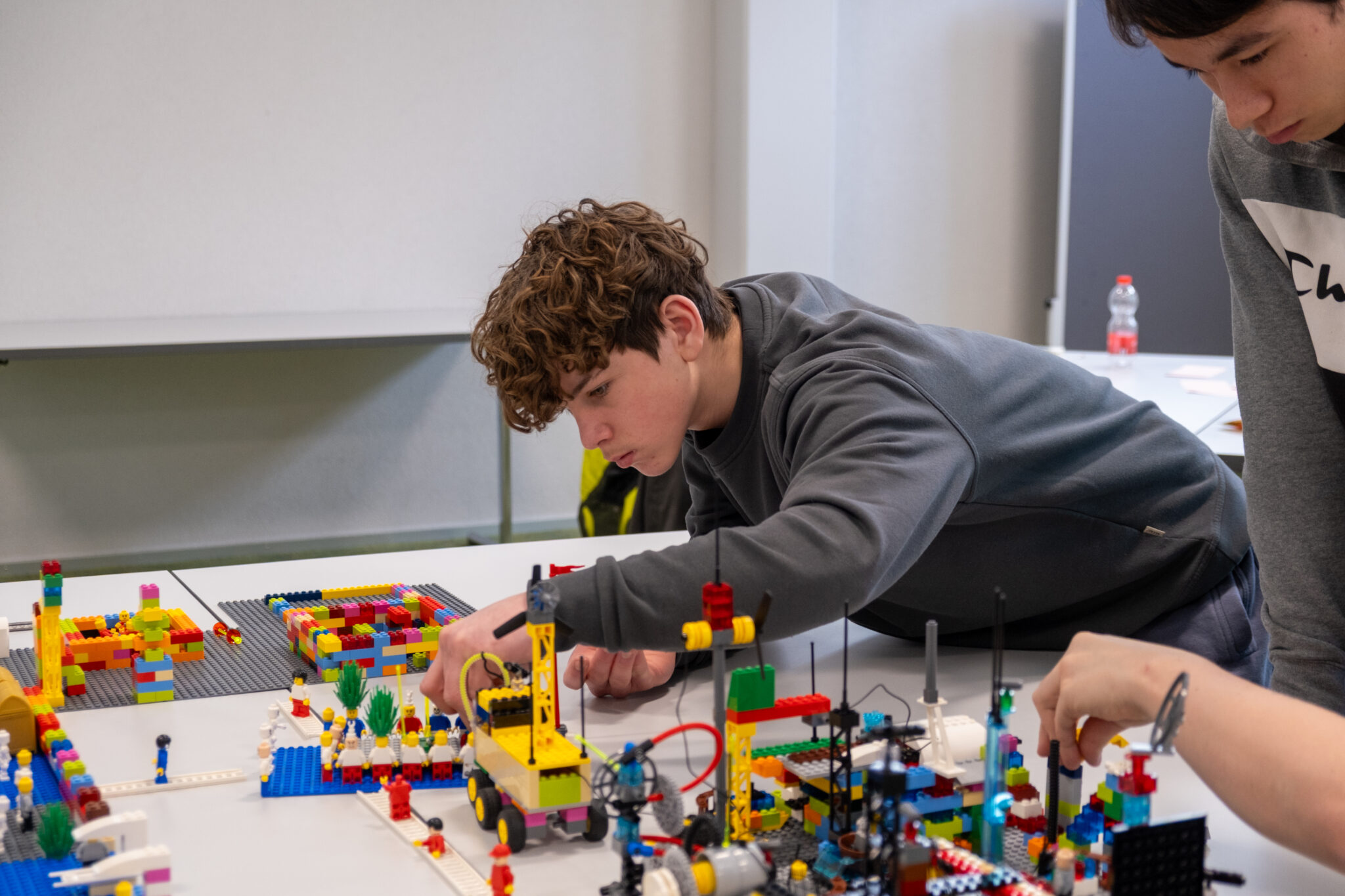 Zwei Teenager bauen auf einem weißen Tisch bunte LEGO-Konstruktionen. Ein Junge mit lockigem Haar beugt sich vor, um ein Teil zu platzieren, während ein anderer daneben steht; beide sind ganz auf ihre Kreationen konzentriert.