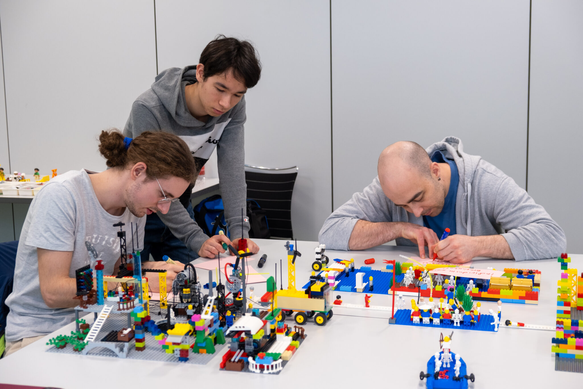 Drei Personen bauen an einem Tisch bunte LEGO-Konstruktionen. Zwei sitzen und konzentrieren sich darauf, die Teile zusammenzusetzen, während eine Person daneben steht und zusieht. Der Tisch ist mit verschiedenen LEGO-Modellen und -Teilen bedeckt.
