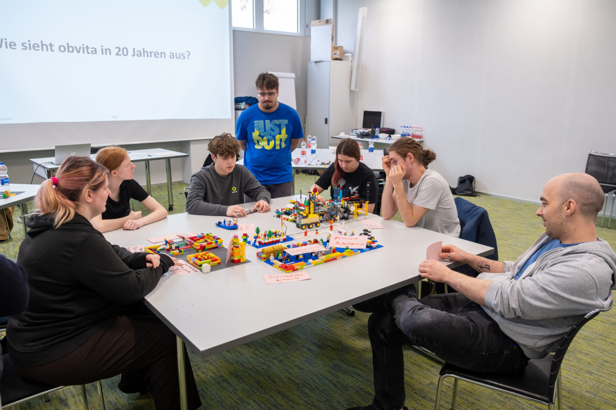 Sechs Personen sitzen mit bunten LEGO-Modellen um einen Tisch herum und nehmen an einer Gruppenaktivität teil. Auf einer Leinwand im Hintergrund wird ein deutscher Text angezeigt. Der Raum ist hell und verfügt über große Fenster.