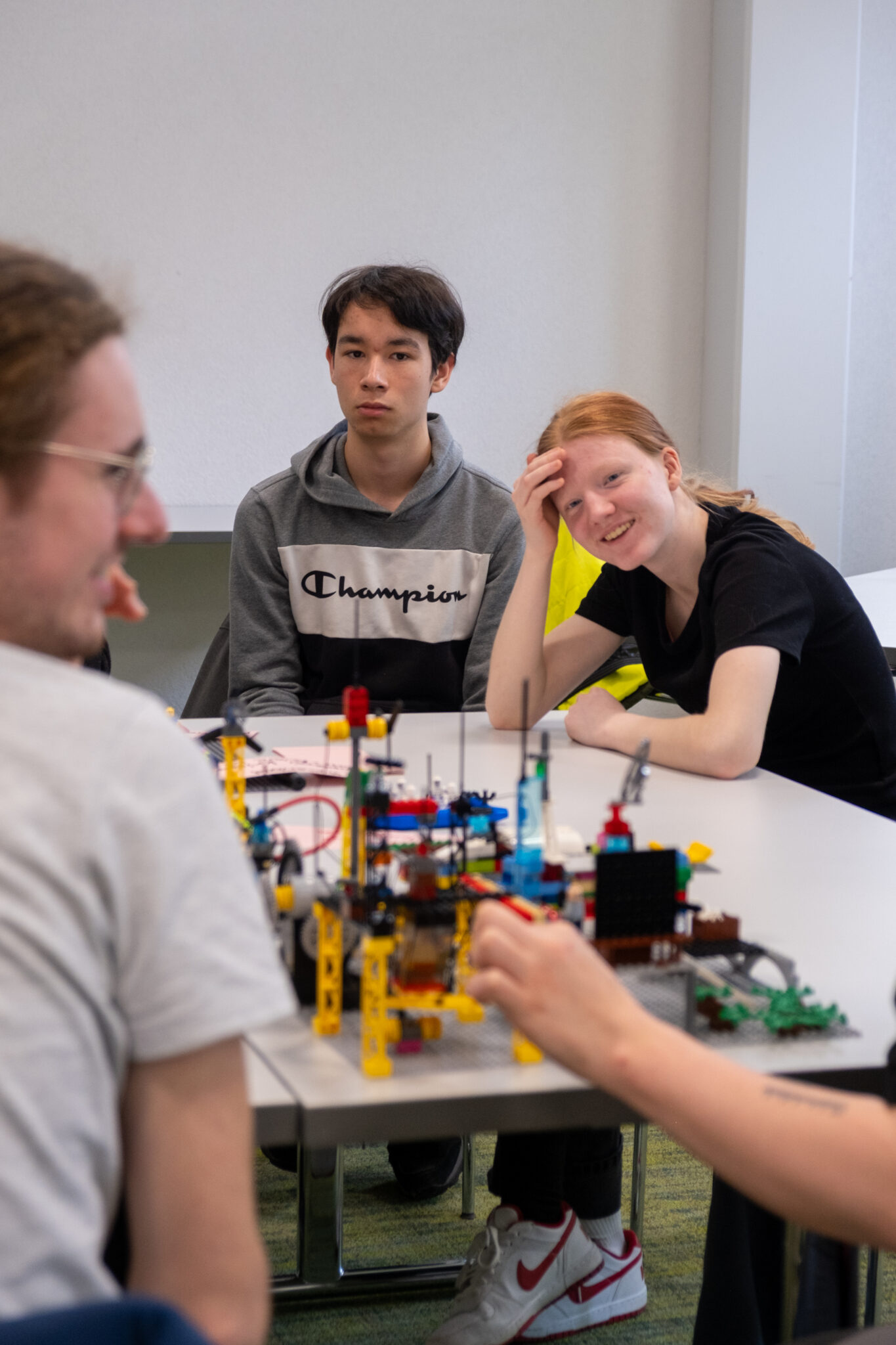Vier junge Leute sitzen um einen Tisch herum, auf dem ein buntes LEGO-Bauwerk steht, und unterhalten sich. Ein Mädchen lächelt und stützt den Kopf auf die Hand, während ein Junge in einem grauen Kapuzenpulli die Gruppe aufmerksam beobachtet.