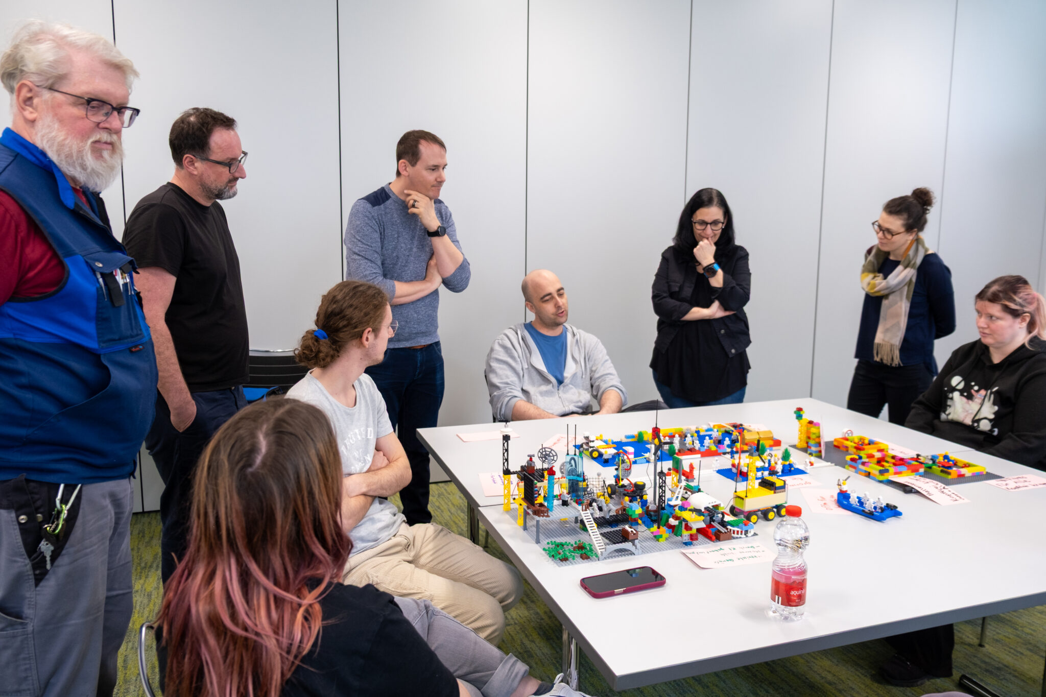 Eine Gruppe von acht Personen versammelt sich um einen Tisch, der mit bunten LEGO-Konstruktionen bedeckt ist, und diskutiert angeregt. Einige sitzen, andere stehen; alle wirken konzentriert und nachdenklich in einem hellen, modernen Raum.
