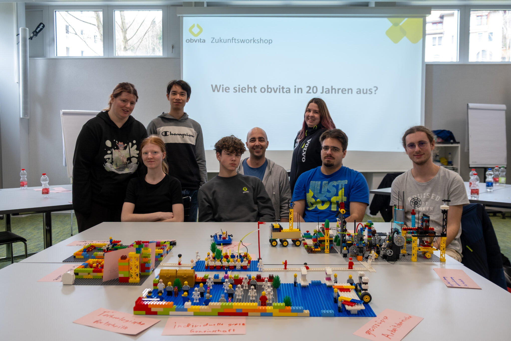 Eine Gruppe von acht Personen posiert hinter Tischen mit bunten LEGO-Kreationen. Hinter ihnen ist auf einer Leinwand „obvita Zukunftsworkshop“ zu sehen sowie die Frage „Wie sieht obvita in 20 Jahren aus?“ auf Deutsch.