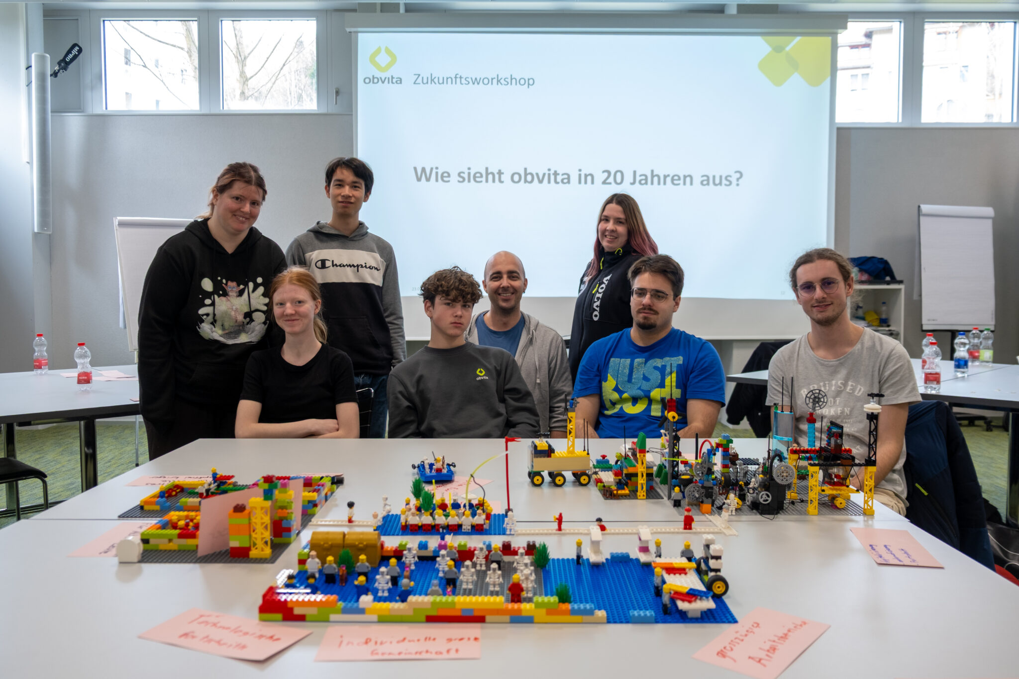 Eine Gruppe von sieben Personen posiert und lächelt vor bunten LEGO-Modellen, die auf Tischen aufgestellt sind. Im Hintergrund ist auf einer Leinwand in einem hellen, modernen Besprechungsraum der Text „Wie sieht obvita in 20 Jahren aus?“ zu sehen.