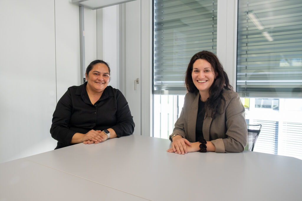 Zwei Frauen sitzen an einem weißen Tisch in einem modernen Büro und lächeln in die Kamera. Beide tragen Geschäftskleidung, und im Hintergrund sind große Fenster mit geschlossenen Jalousien zu sehen.