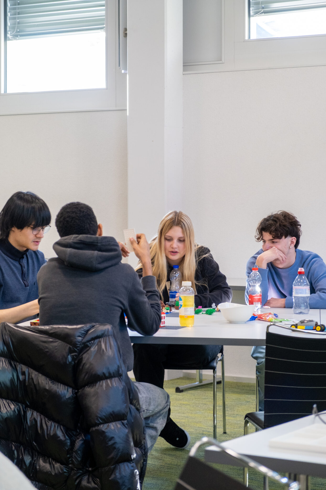 Vier Schüler sitzen in einem hellen Klassenzimmer um einen Tisch herum und unterhalten sich; auf dem Tisch liegen Snacks, Wasserflaschen und Schreibutensilien.